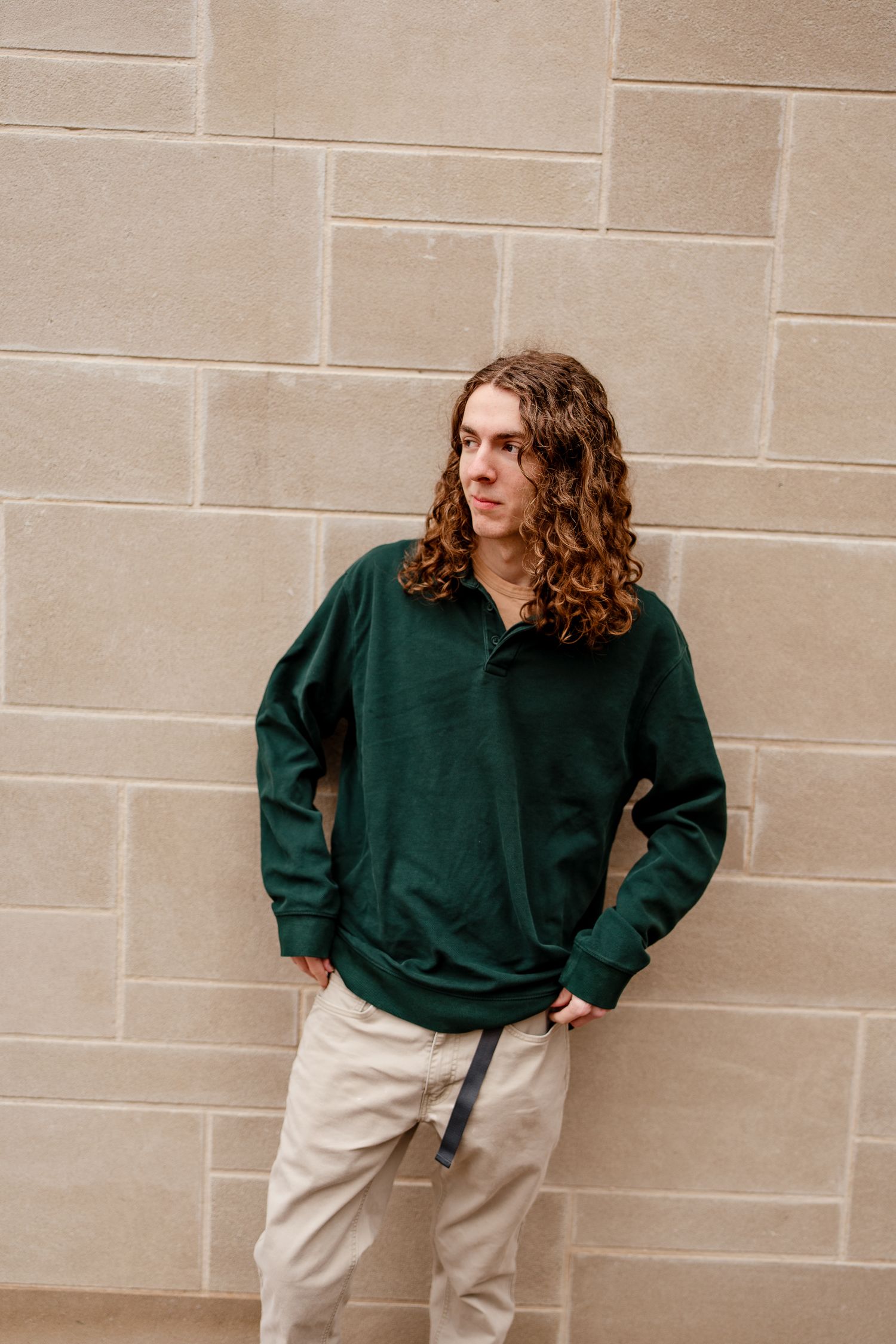 Person in dark green sweater and khakis posing against beige brick wall.