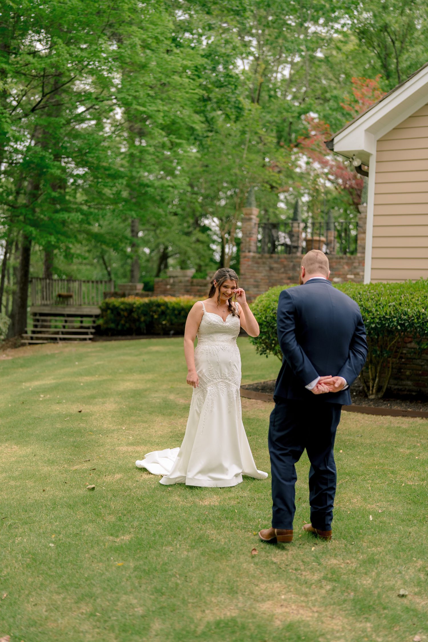 Emotional bride in white gown wiping tears during first look with groom in navy suit on green lawn.