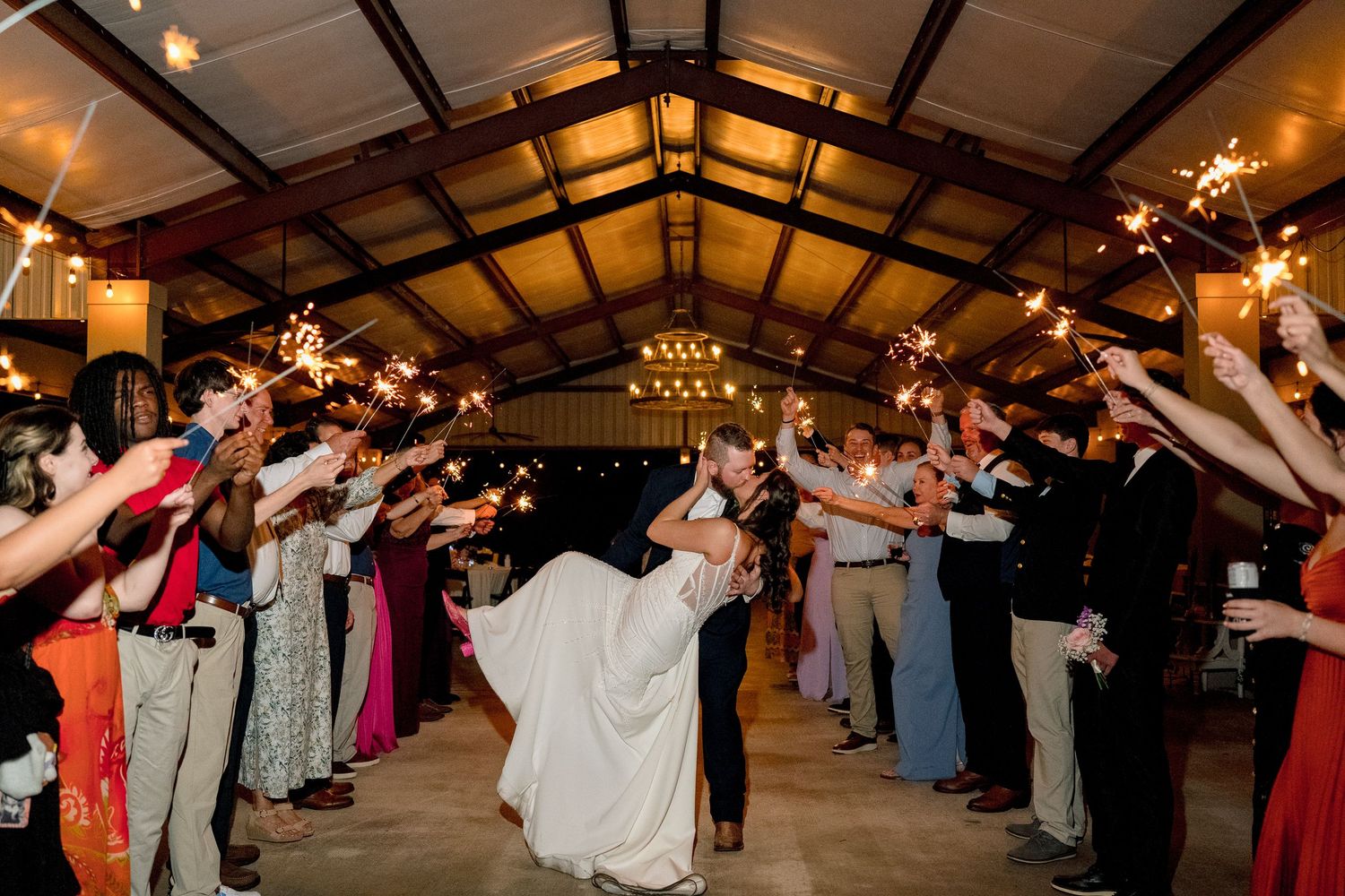 Bride and groom exit wedding reception through sparkler tunnel surrounded by cheering guests indoors.