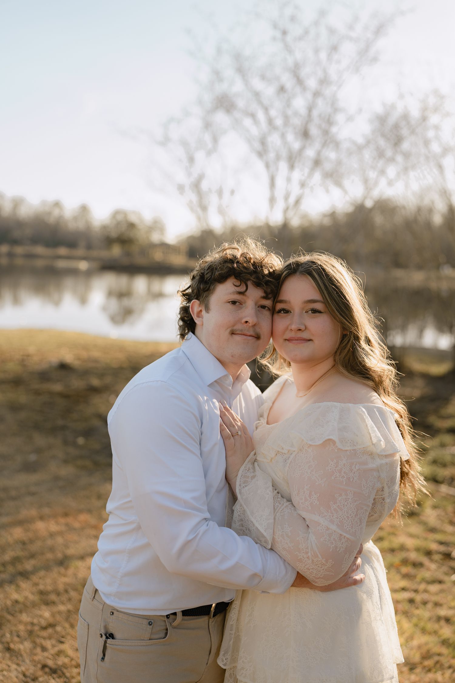 A young couple embraces near a serene lake at golden hour, dressed in elegant white and cream attire.