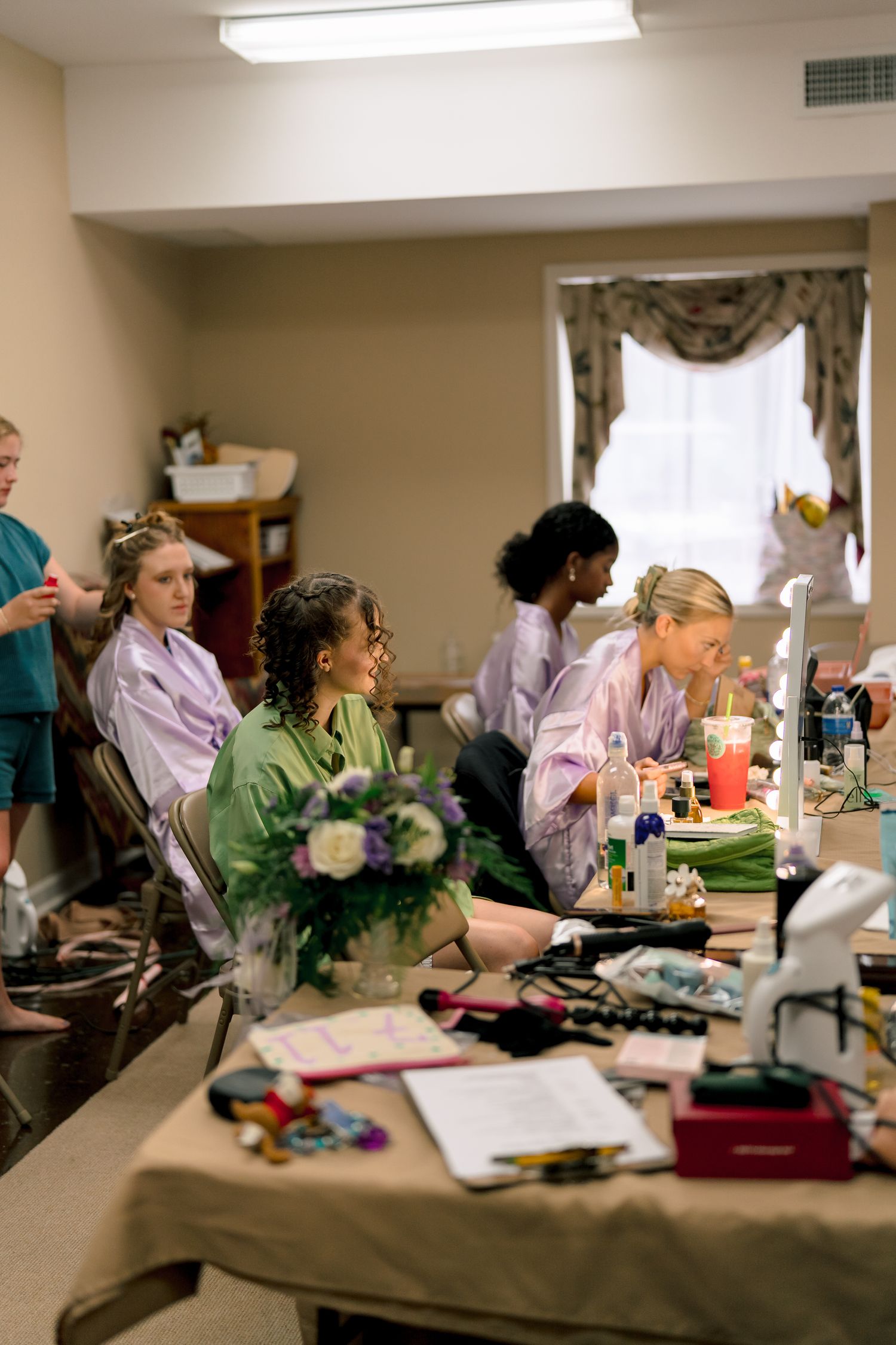 Bridesmaids in pastel robes sit at a makeup table getting ready for a wedding ceremony.