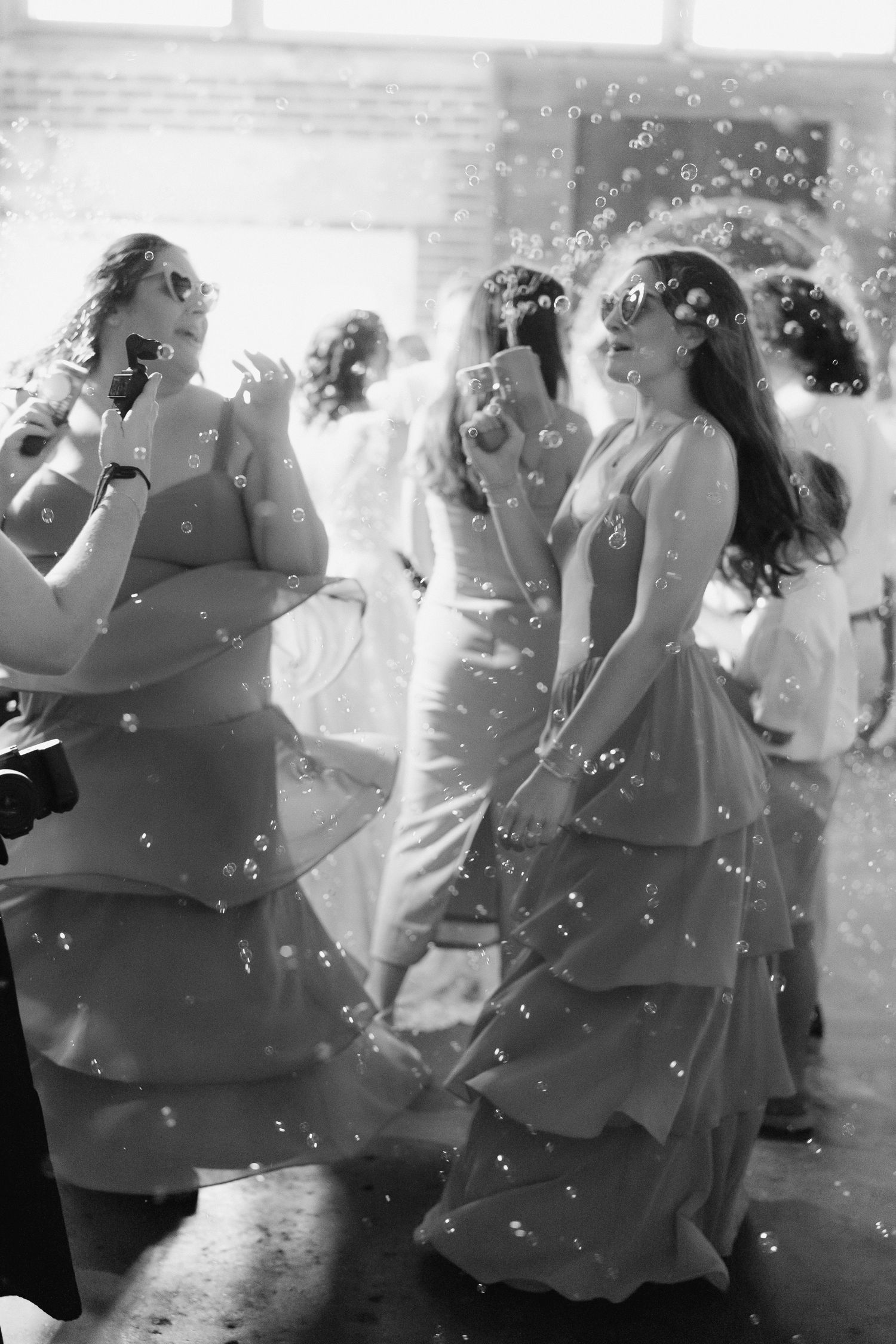 Bridesmaids in elegant gowns dancing and blowing bubbles at a wedding reception, black and white.