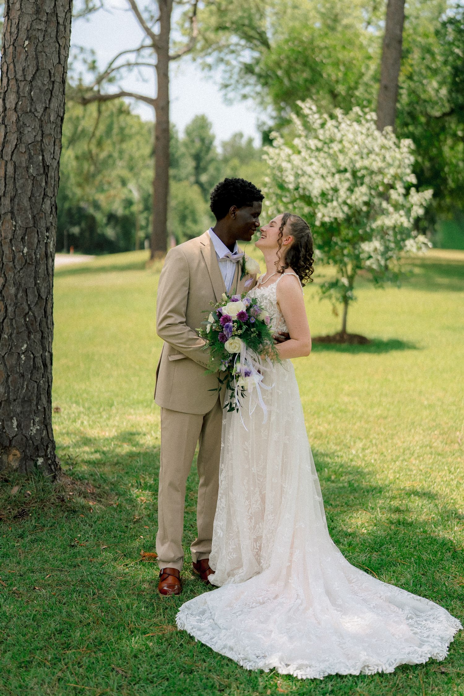 Interracial couple sharing a kiss outdoors in lush garden, bride in lace gown with bouquet.