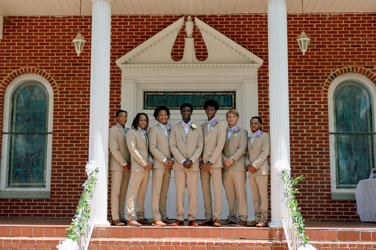 Groom and groomsmen in tan suits posing on brick church steps before wedding ceremony.