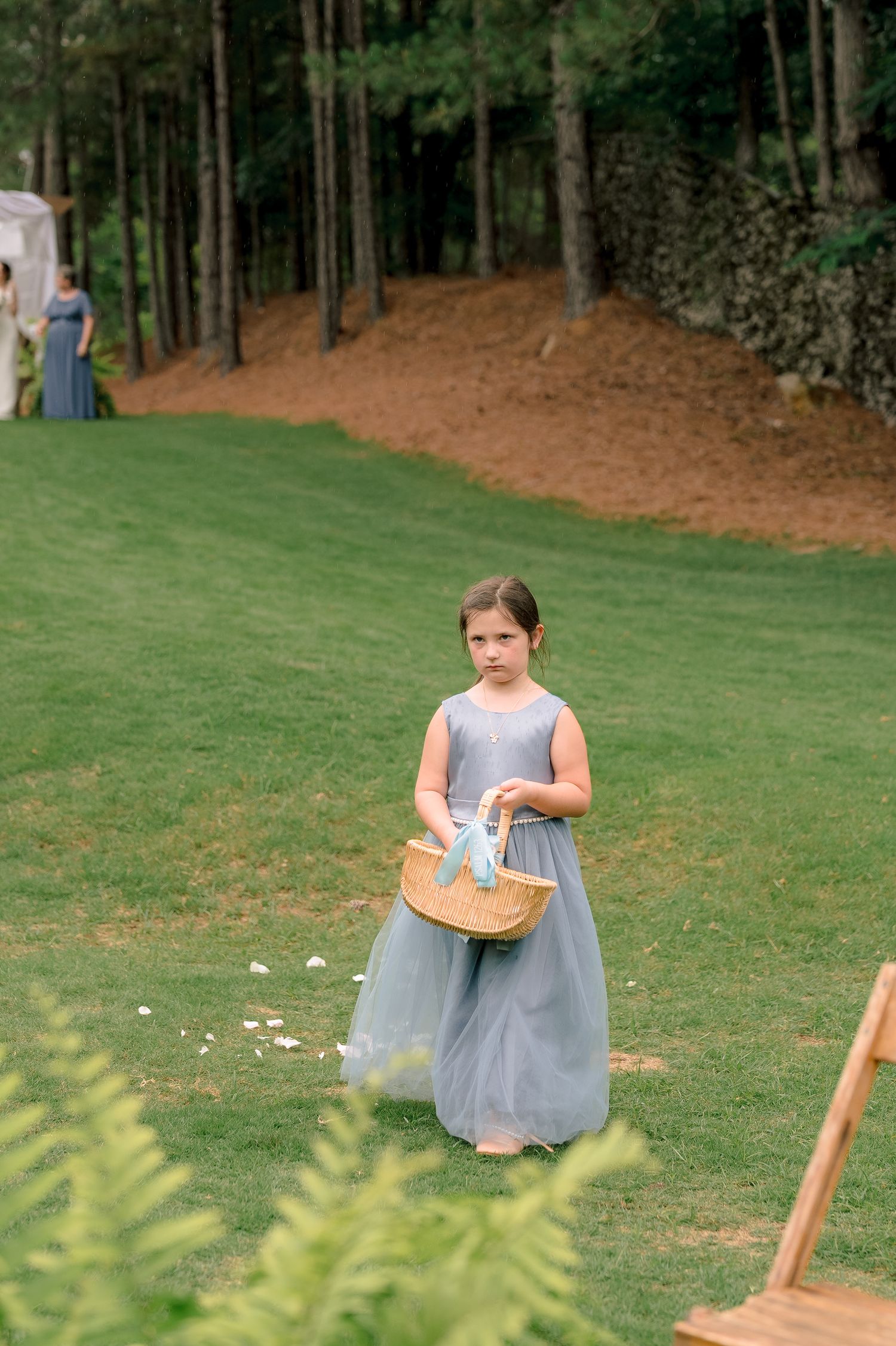 Young flower girl in dusty blue dress carrying a wicker basket on a green lawn.
