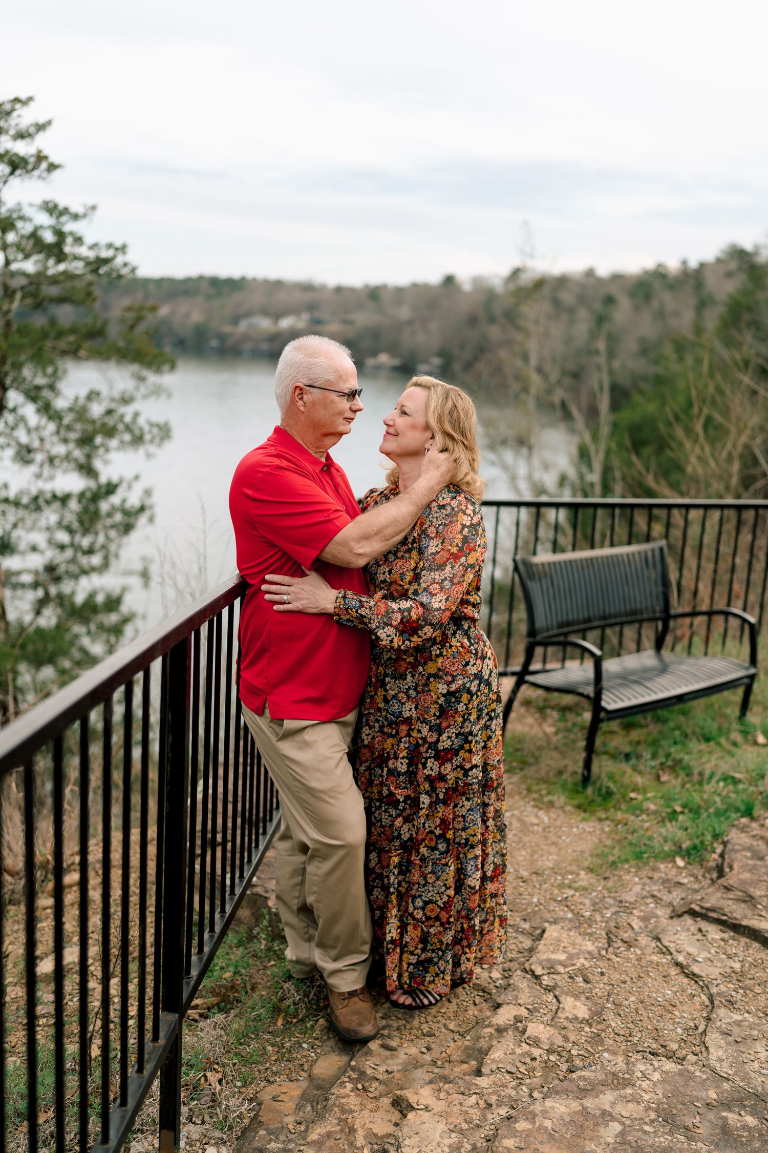 Couple embracing on a scenic overlook with railing, lake, and trees in the background.