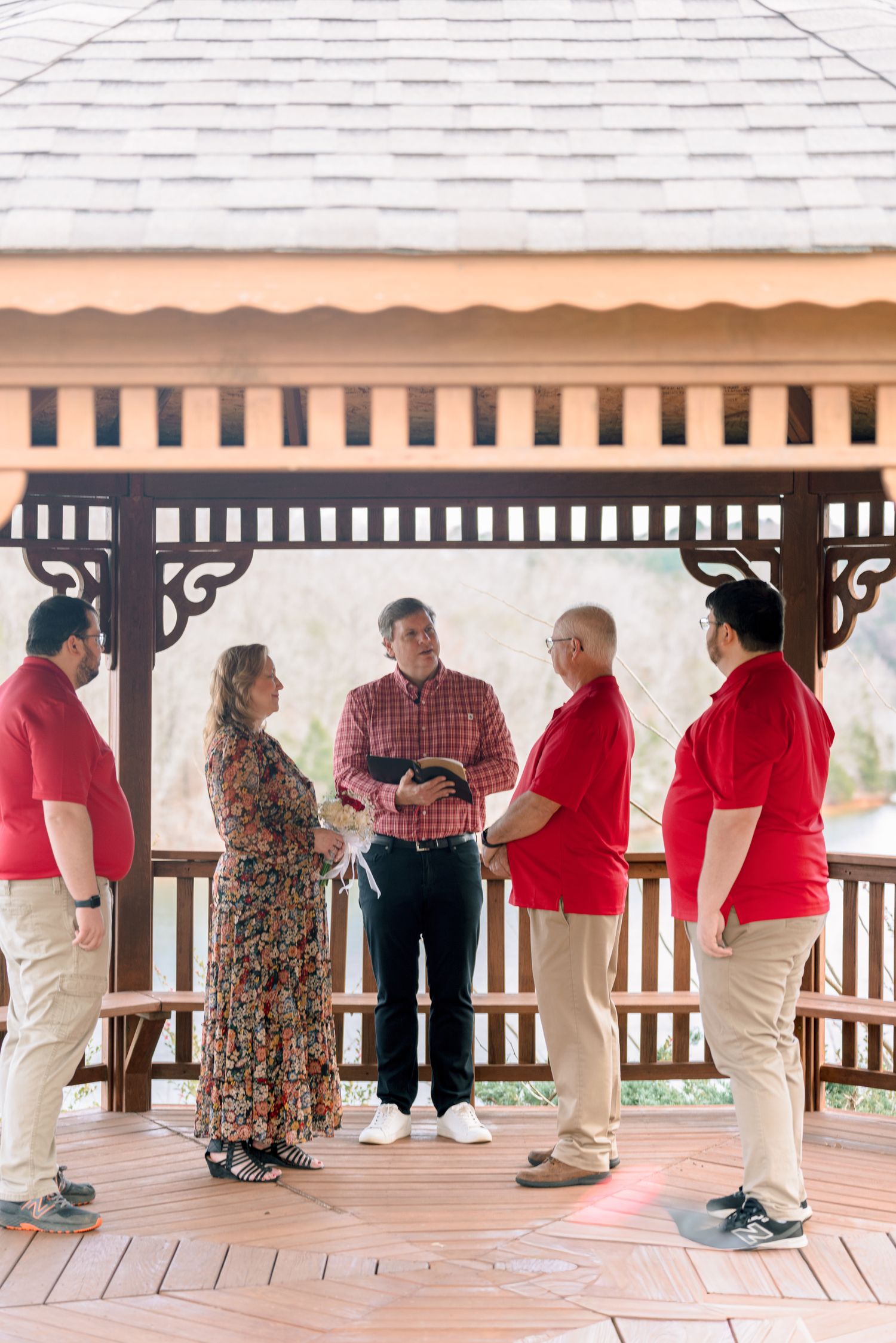 Small wedding ceremony in a gazebo with officiant holding a Bible and guests in red polo shirts.