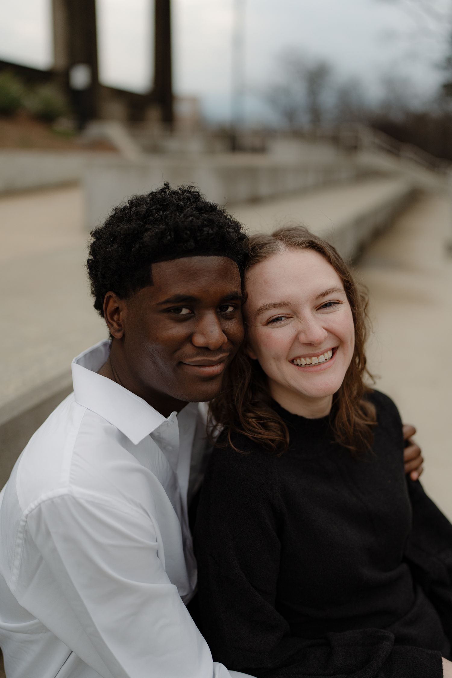 A smiling interracial couple poses closely together outdoors, the woman in black and man in white shirt.