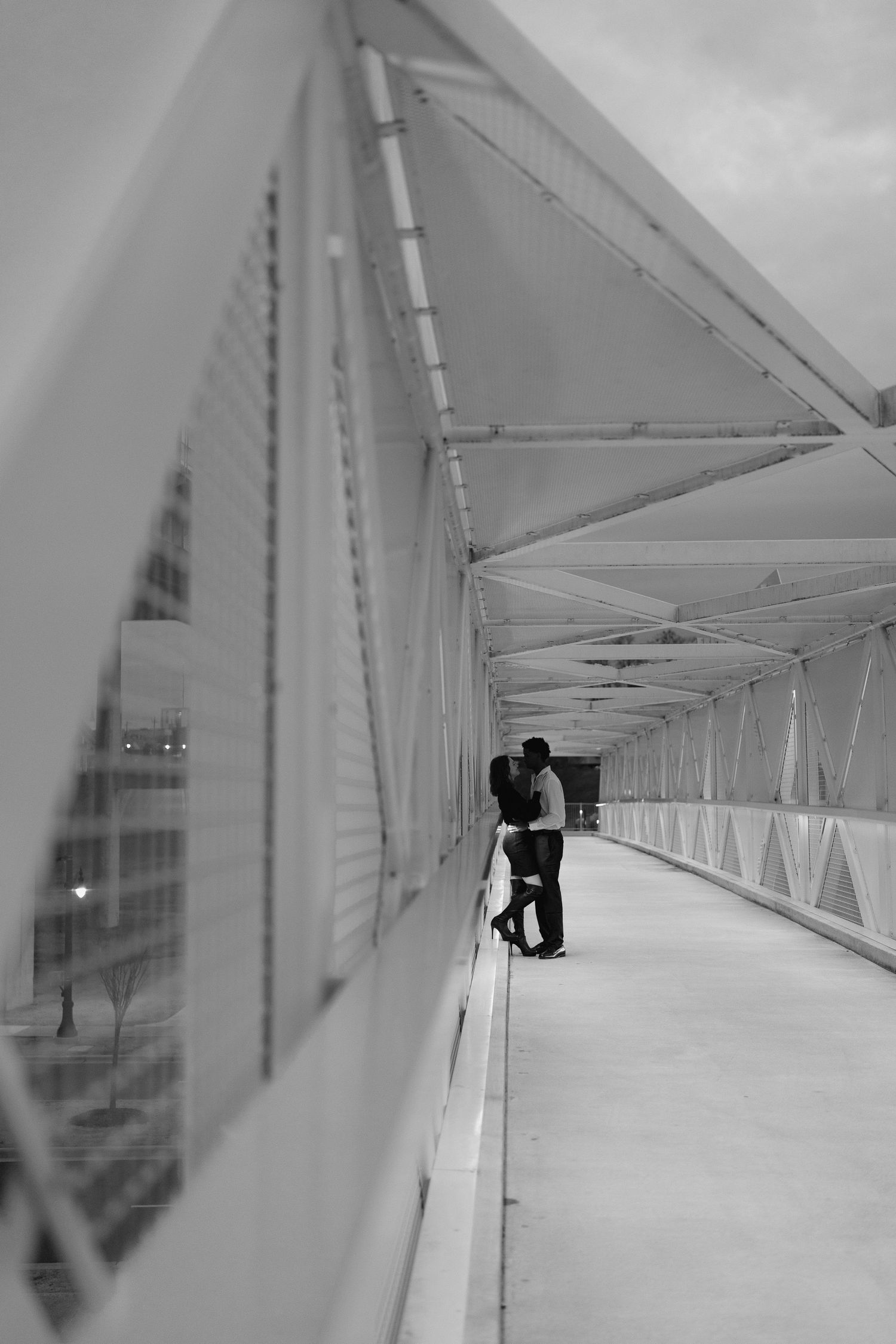 A couple shares a romantic kiss on a dramatic enclosed bridge walkway in black and white.