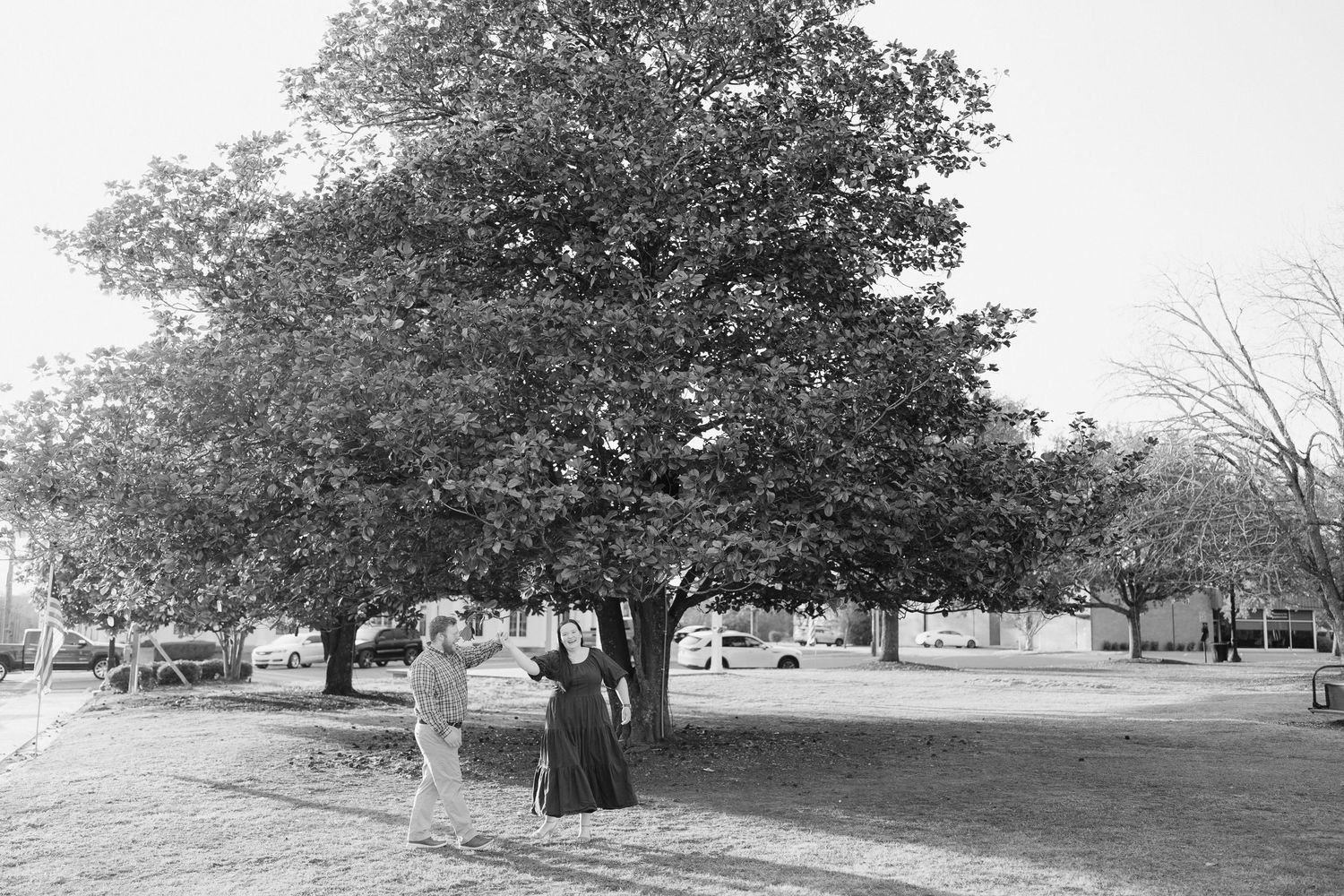 Black and white photo of a couple posing together under a large tree in an open park setting.