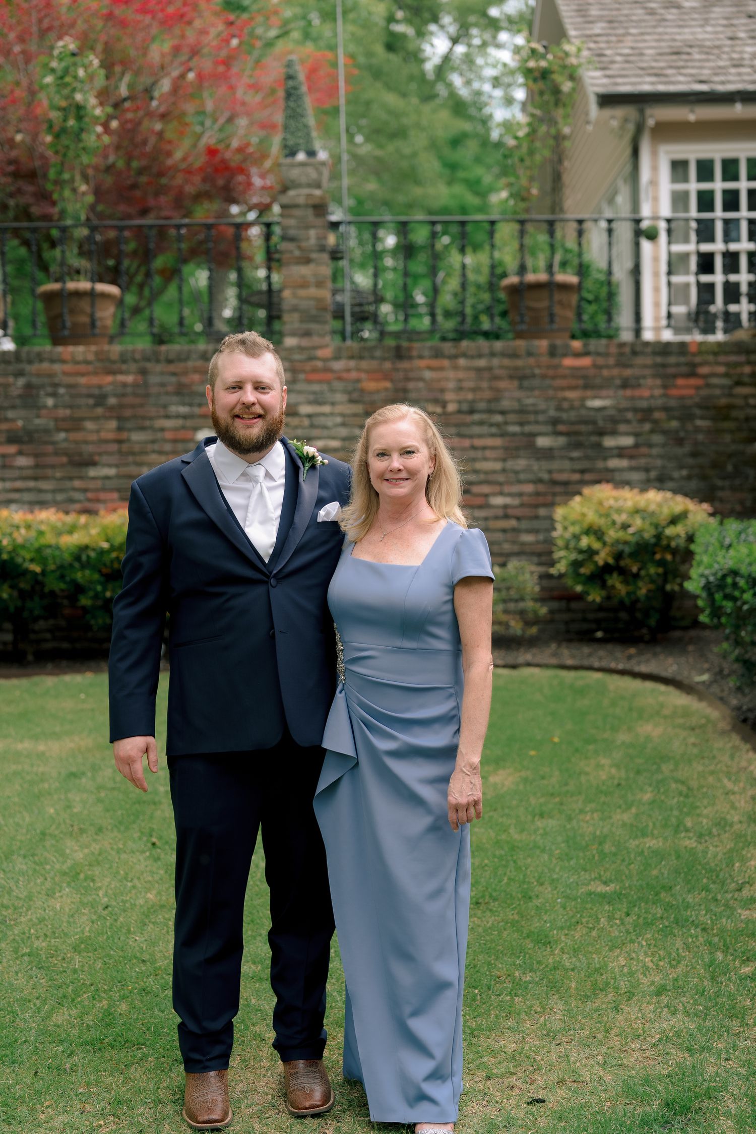 Couple posing outdoors at a wedding venue, man in navy suit, woman in blue gown, brick building background.
