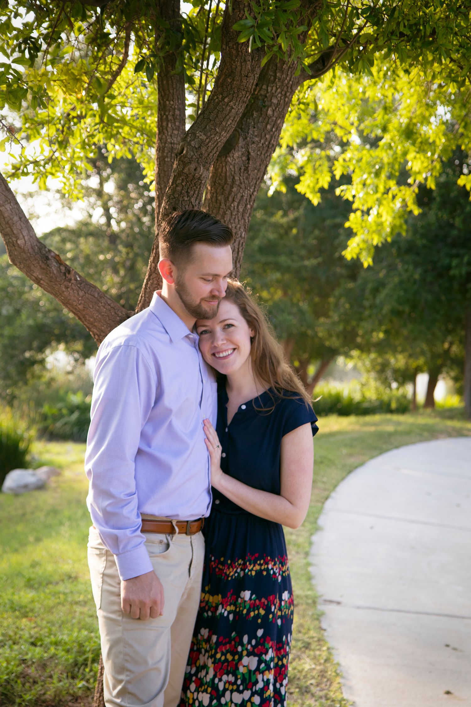 Couple embracing on a sunlit path surrounded by lush green trees.
