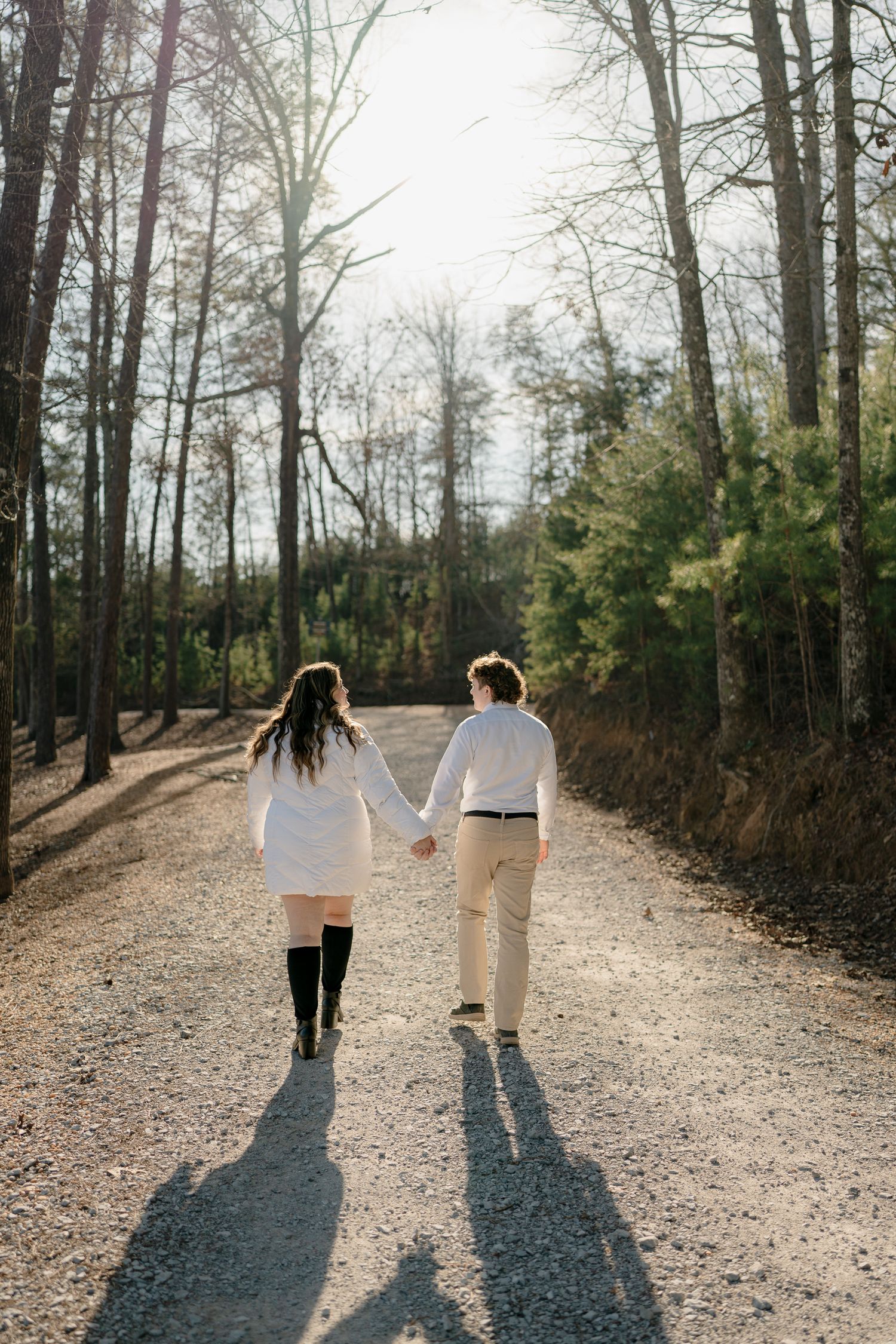 Couple holding hands walking down a sunlit gravel path surrounded by tall trees in a forest.
