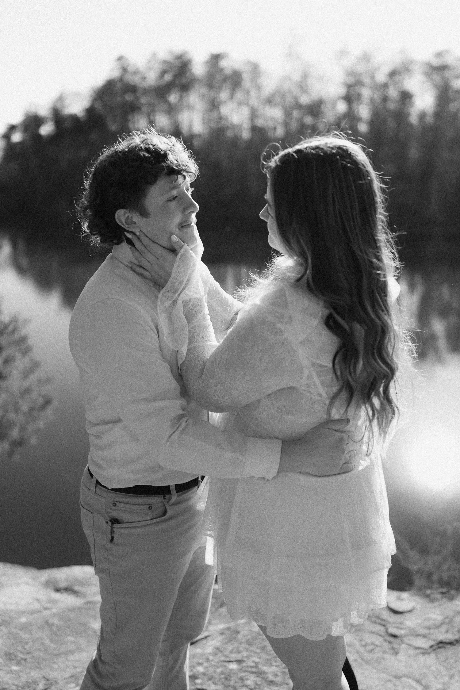Black and white photo of couple gazing at each other tenderly by a reflective lake outdoors.
