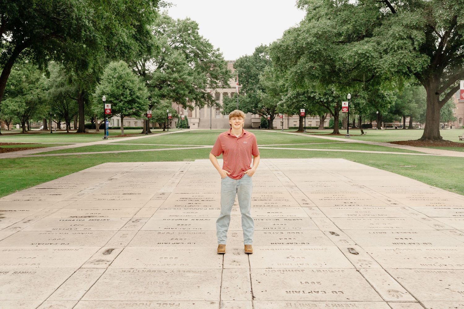 Figure in coral shirt stands centered on a wide campus walkway surrounded by green trees.