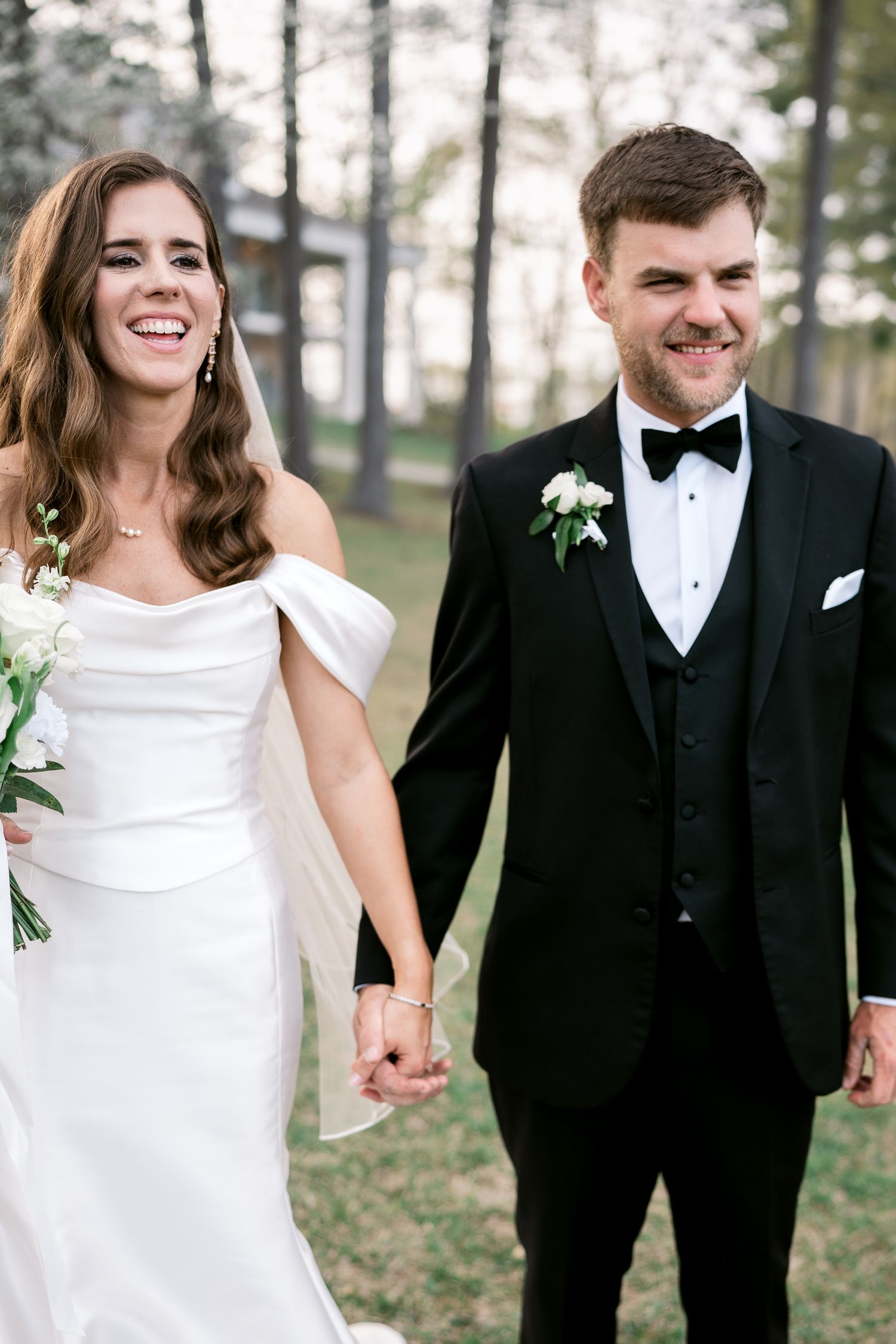 Happy newlyweds hold hands and smile while walking through pine forest.