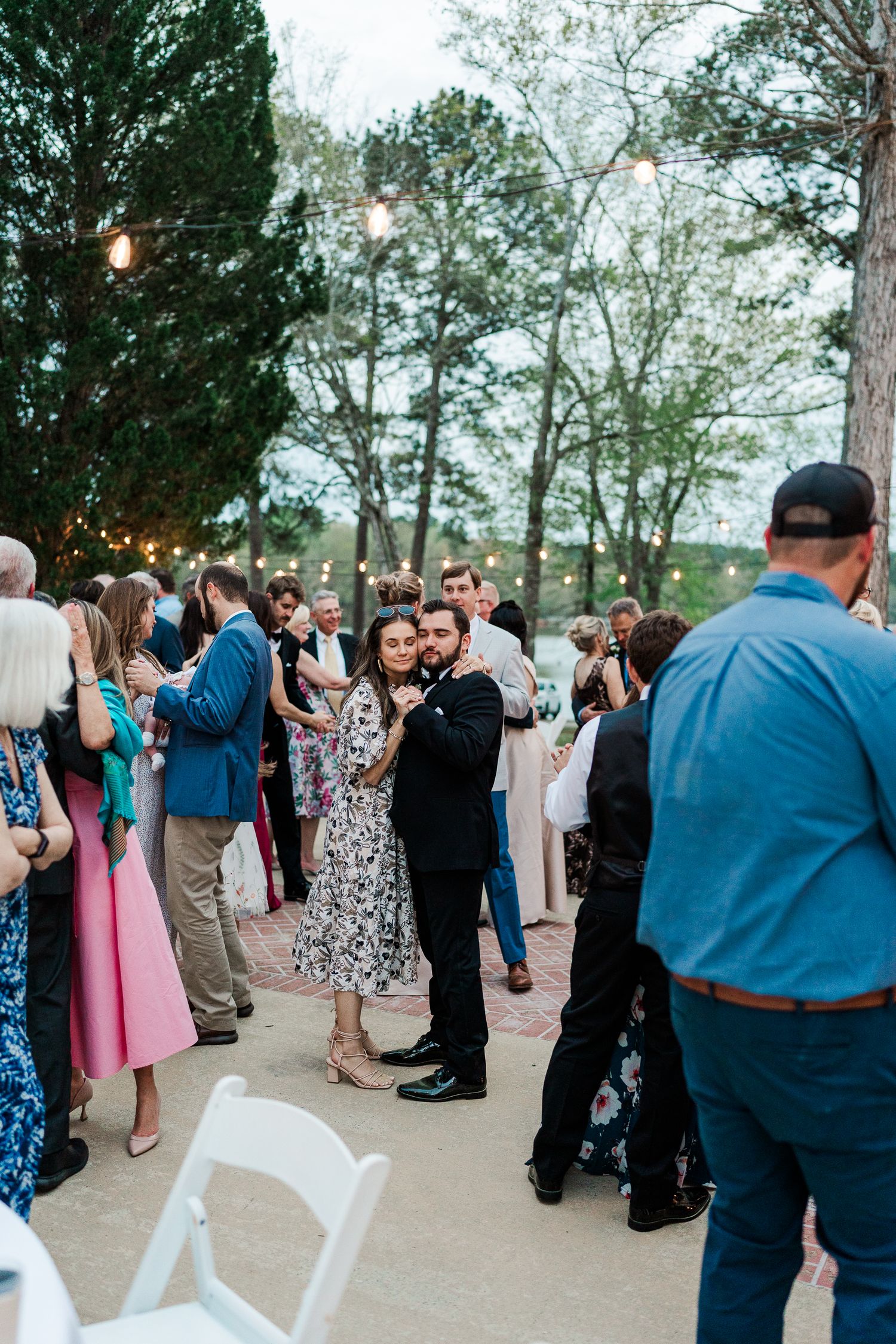 Guests mingle and dance during an outdoor wedding reception surrounded by pine trees at dusk.