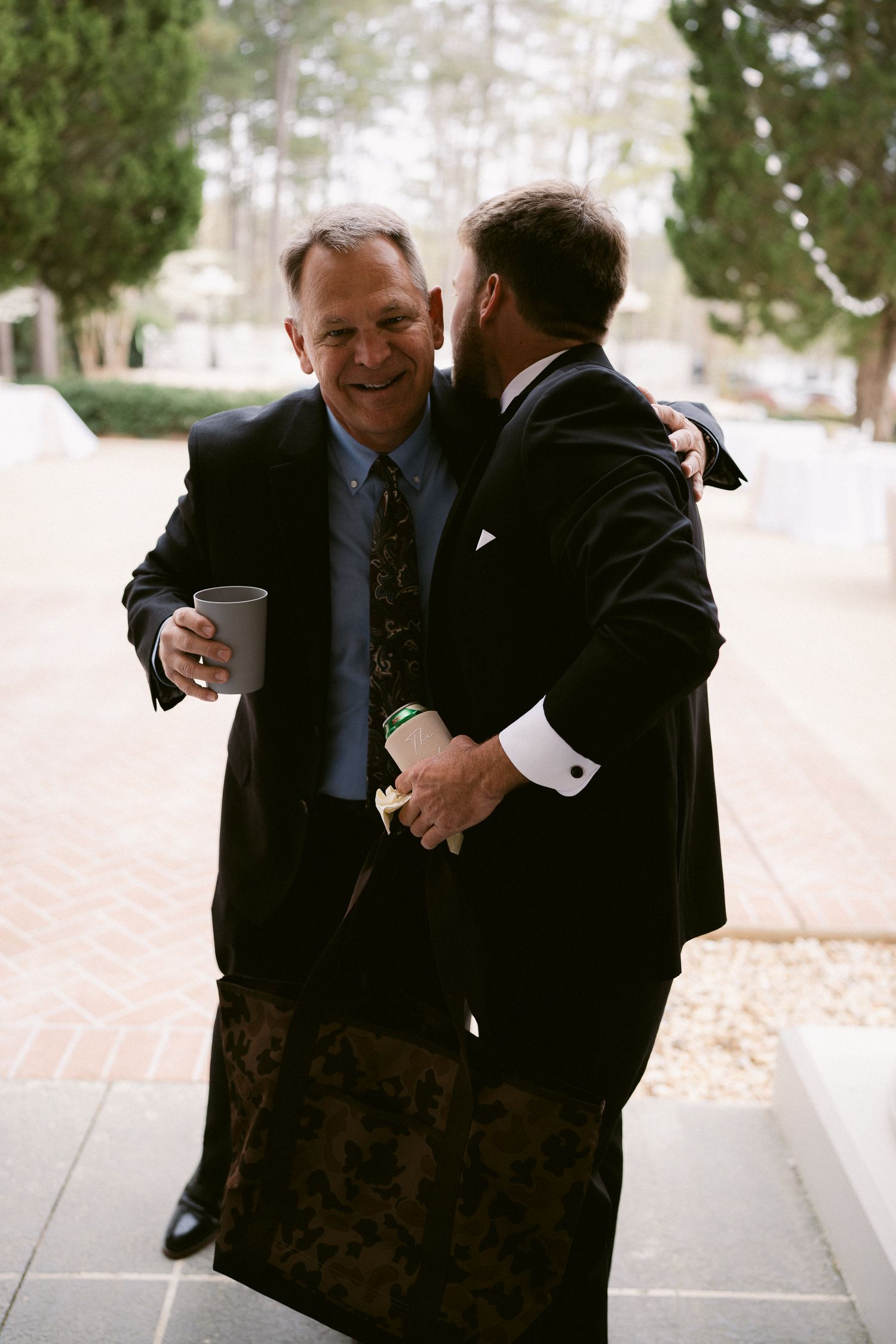 Two men in suits share a lighthearted moment outdoors, with one holding a coffee cup.