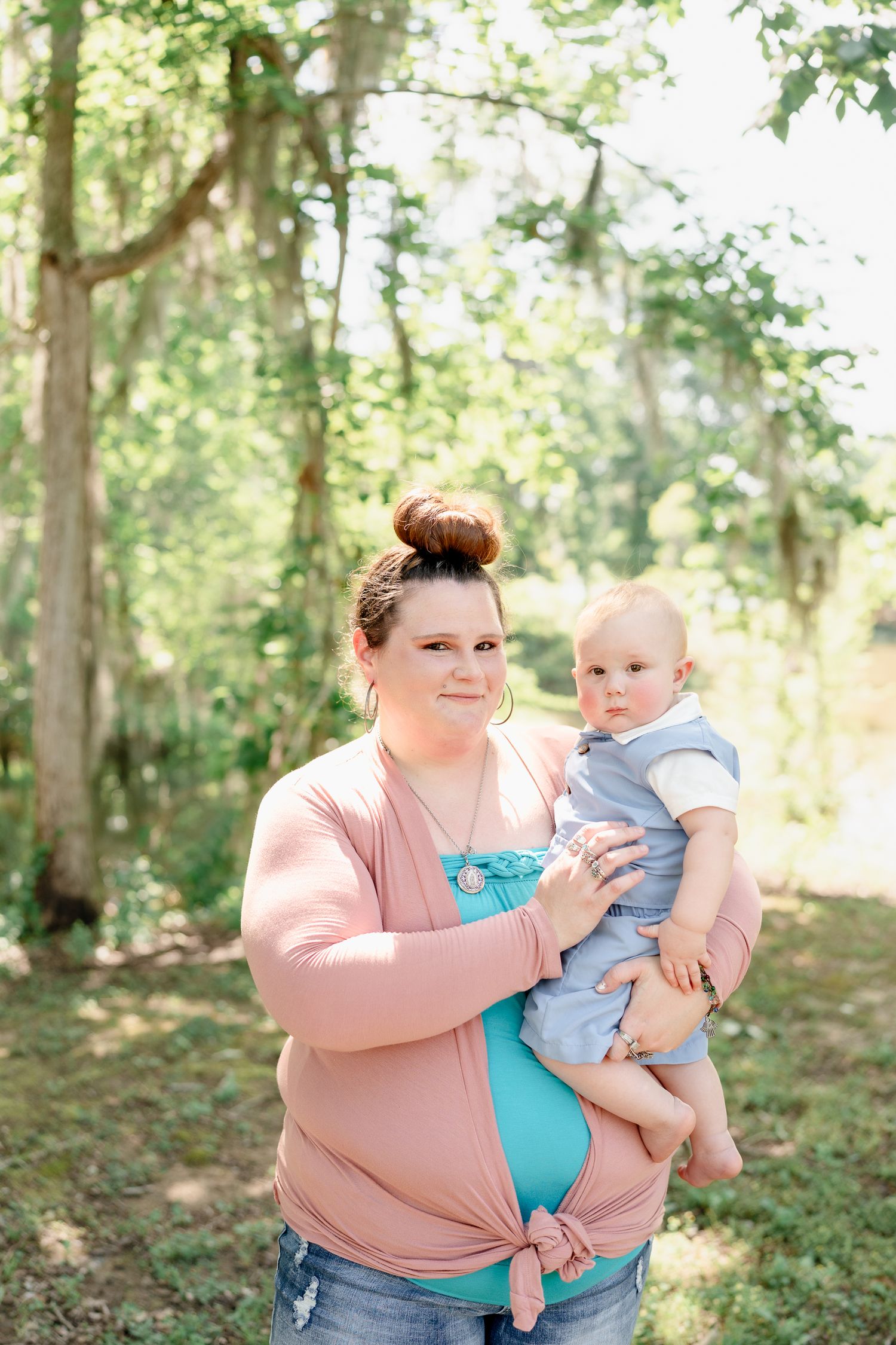Woman in pink sweater holding baby in blue outfit against leafy forest background.