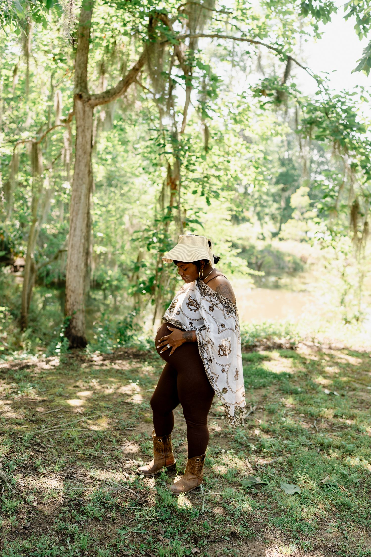Figure in flowing printed dress and hat poses in a sunlit forest setting.