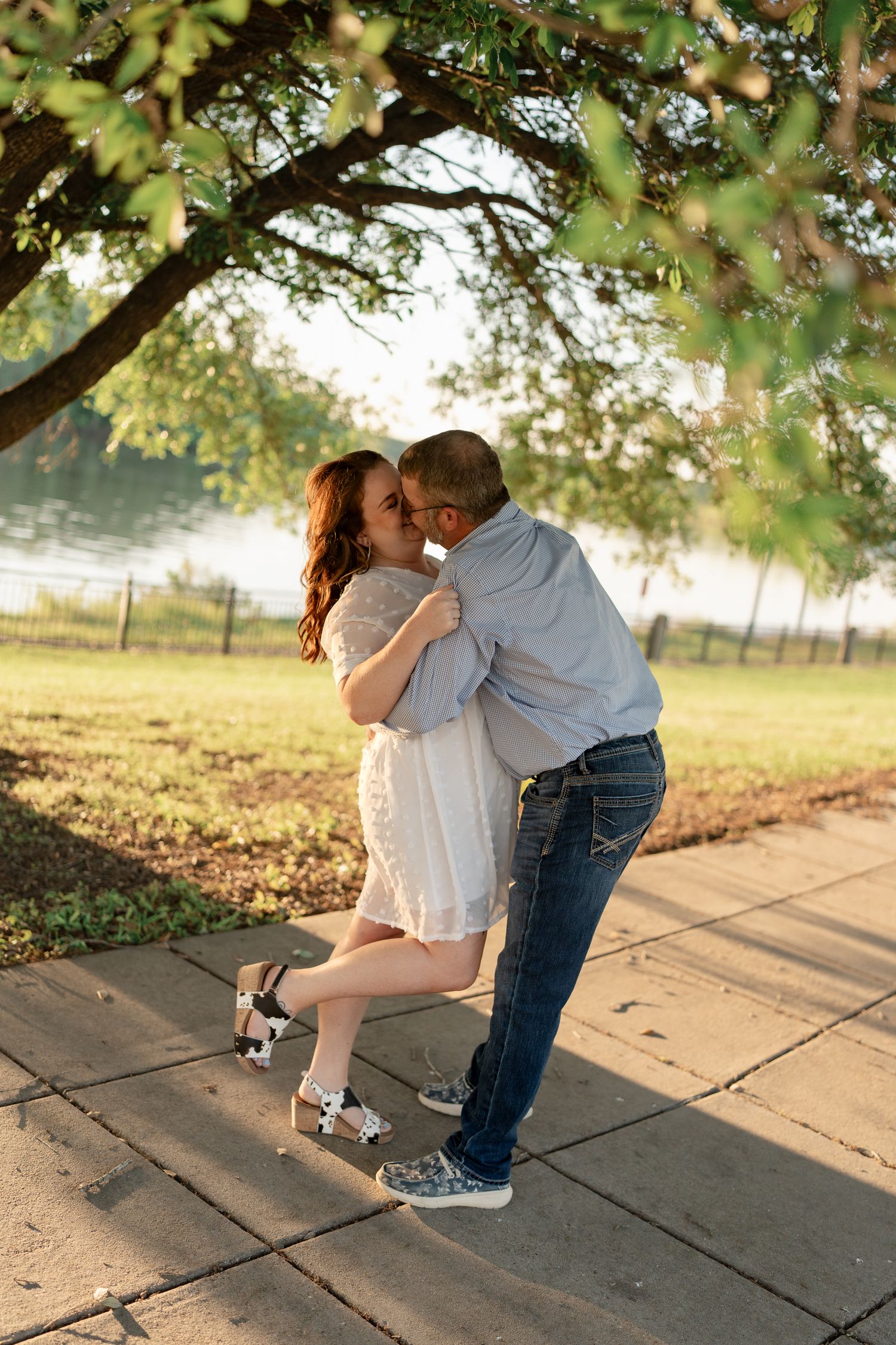 Romantic couple in casual attire dancing together under tree branches at dusk.