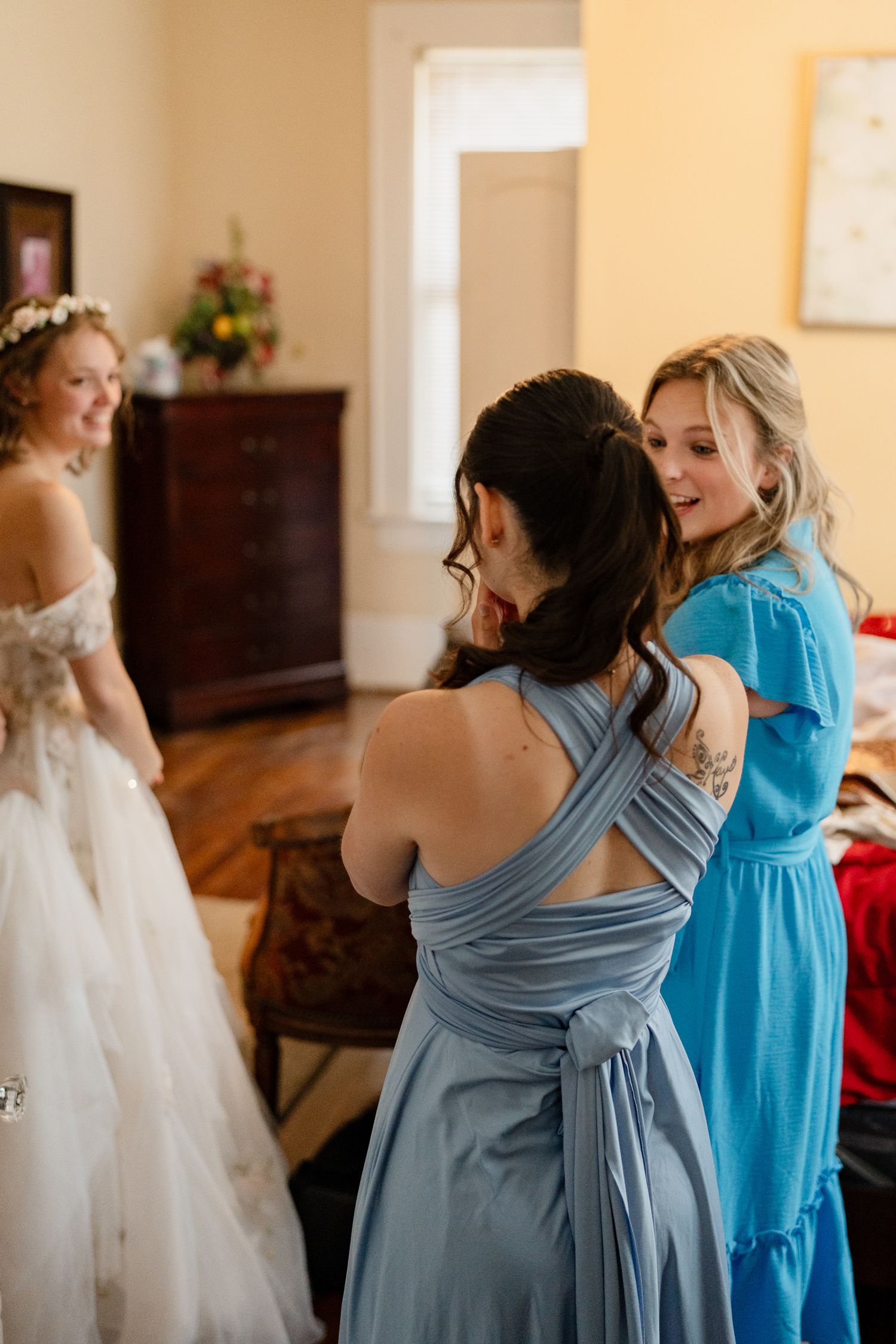 Bridesmaids in blue dresses assisting with wedding preparations in a sunlit room.