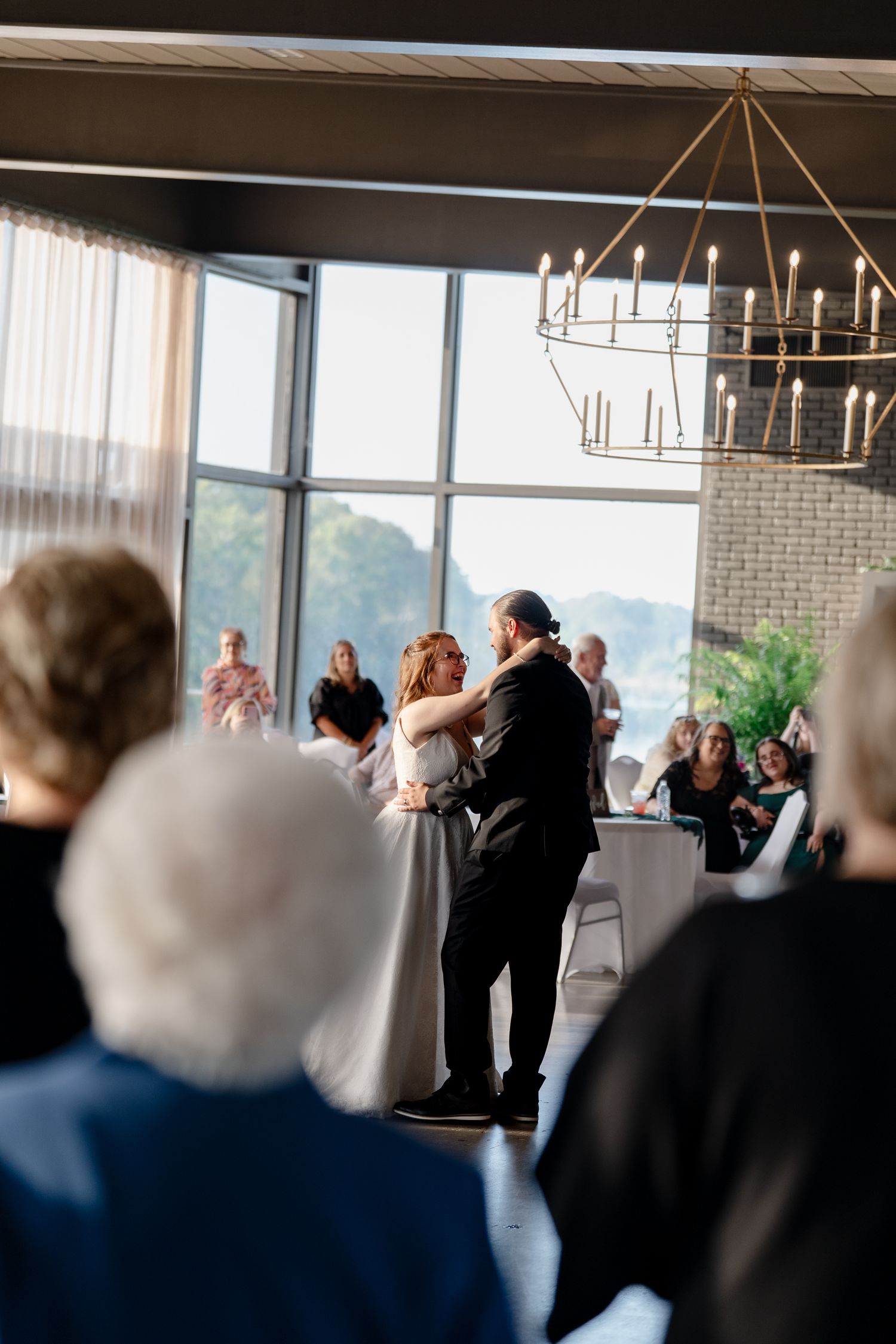 A couple shares their first dance at a wedding reception with a modern chandelier and large windows overlooking nature.