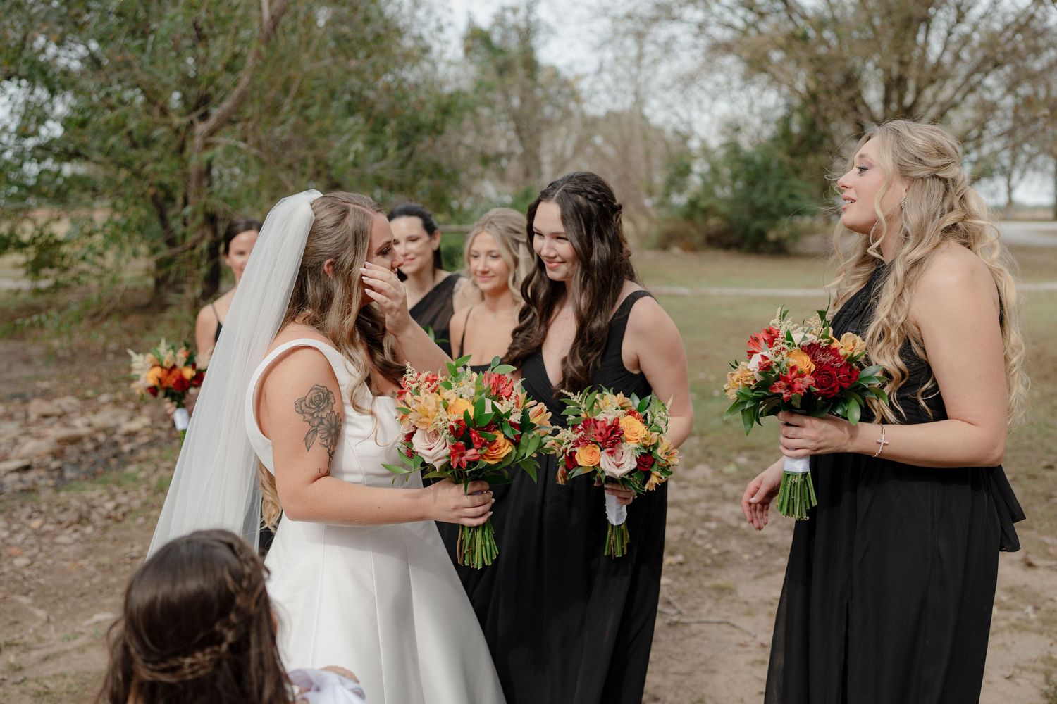 Wedding party members in black dresses share joyful moments with bouquets of vibrant flowers in an outdoor setting.