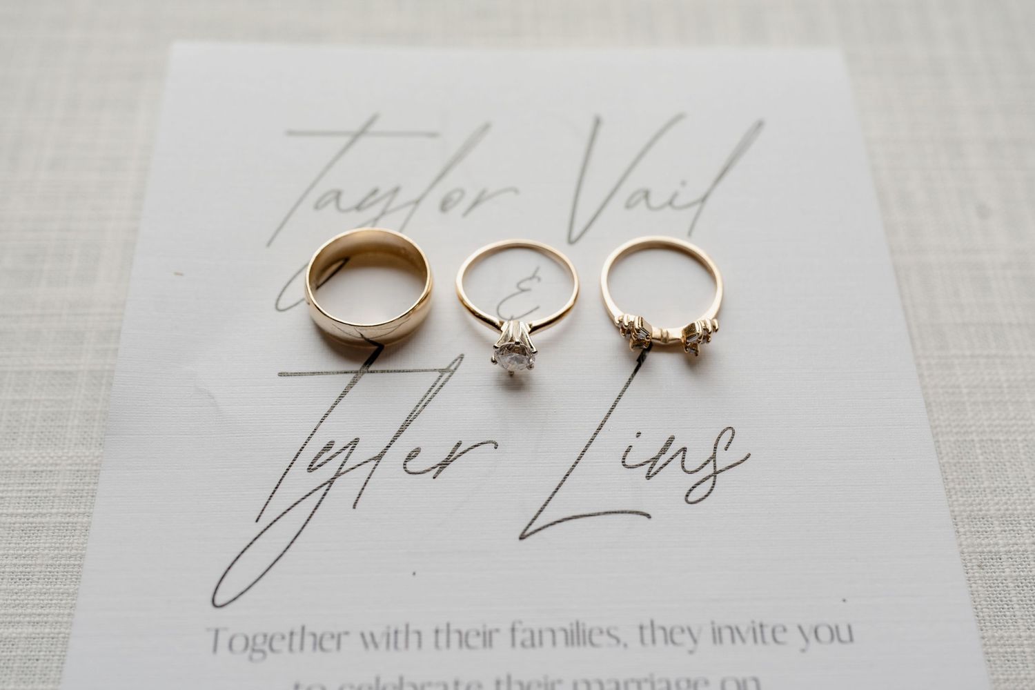 Wedding invitation details and rings displayed on a white textured surface with elegant calligraphy.