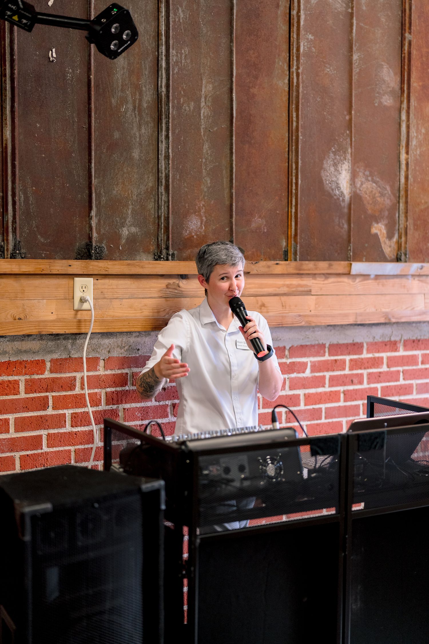Woman with short gray hair speaking into microphone at DJ booth against rustic brick and wood wall backdrop.