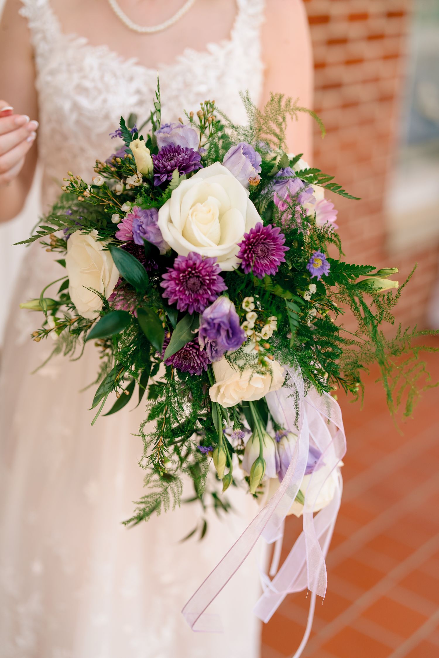 Bride holding lush wedding bouquet with white roses and purple flowers with green foliage.