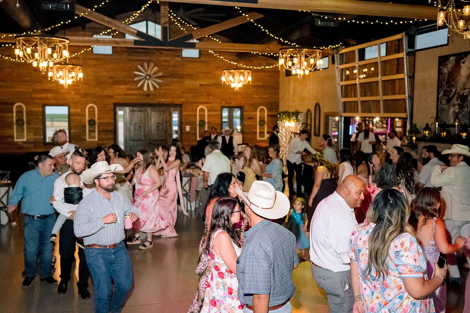 Guests dancing at a rustic western wedding reception with chandeliers and wood walls.