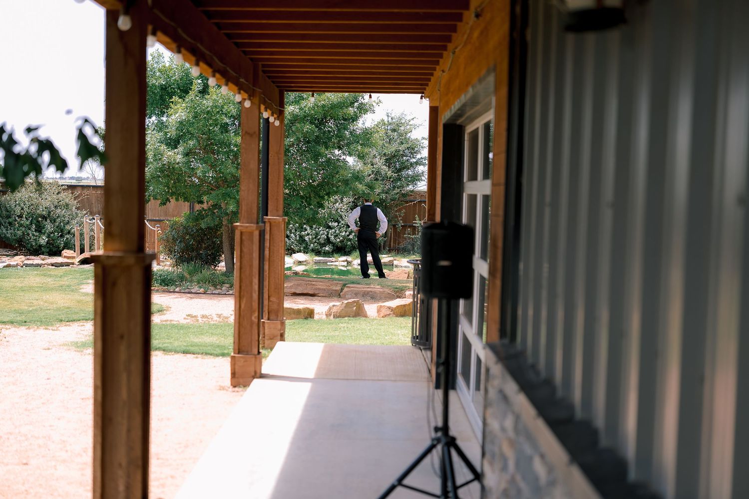 A speaker on a stand sits on a covered patio overlooking a sunny outdoor wedding venue with a guest in the background.
