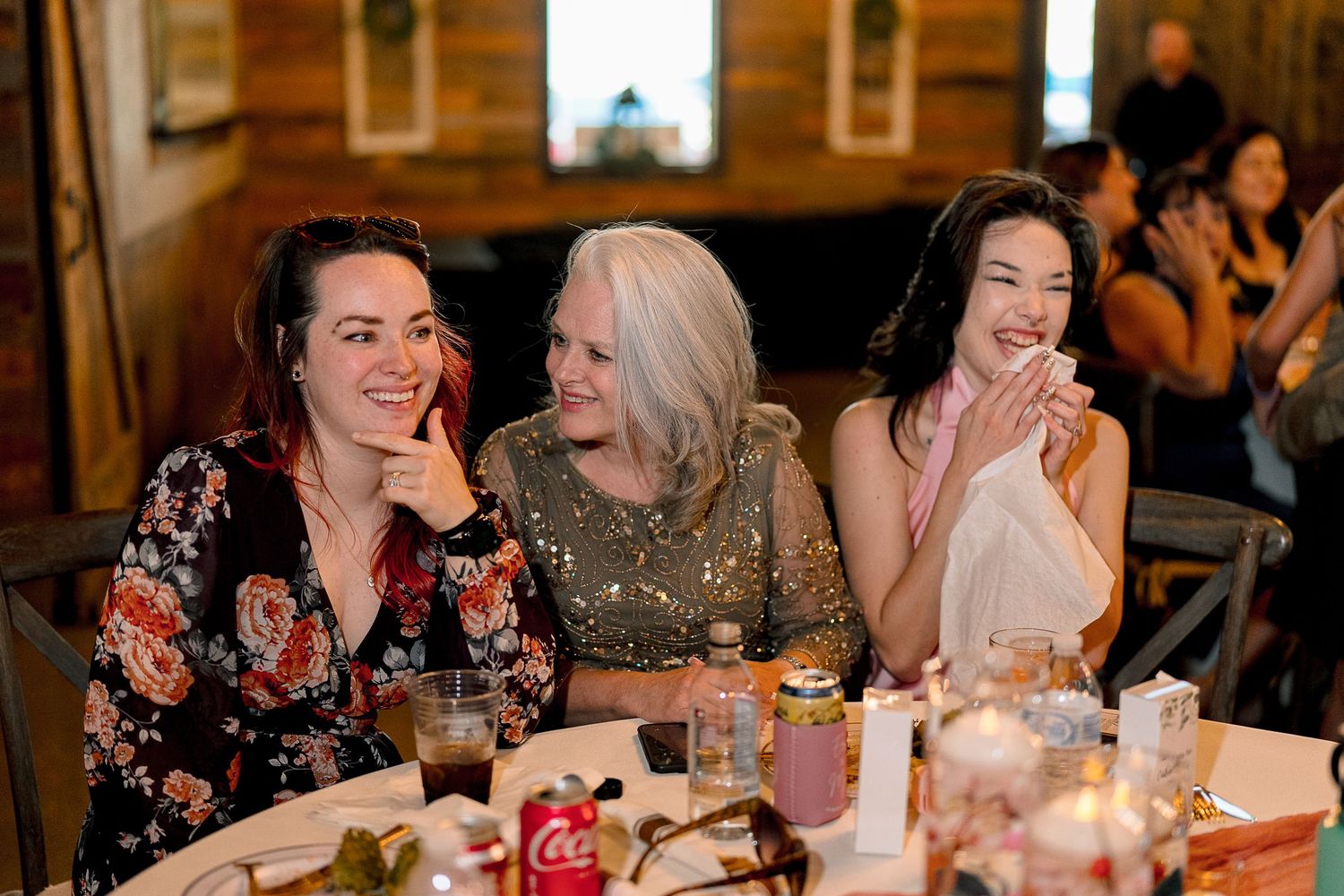Three women laughing and smiling together at a wedding reception dinner table with festive decorations.