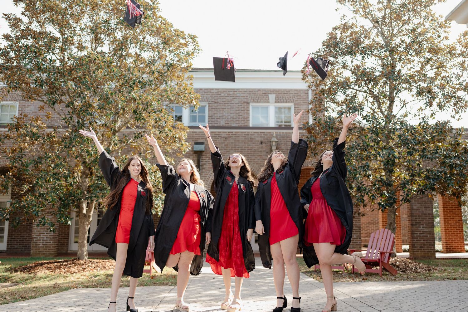 A group in red dresses and black jackets poses together in a line outdoors near a brick building.