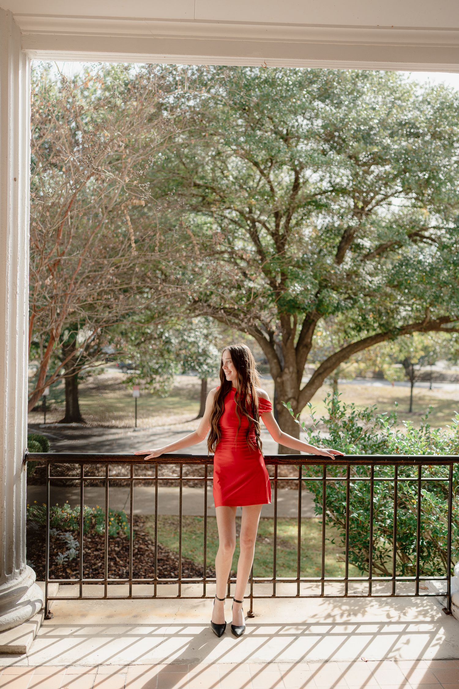 A figure in a red dress stands on a white porch overlooking a scenic landscape with trees in the background.