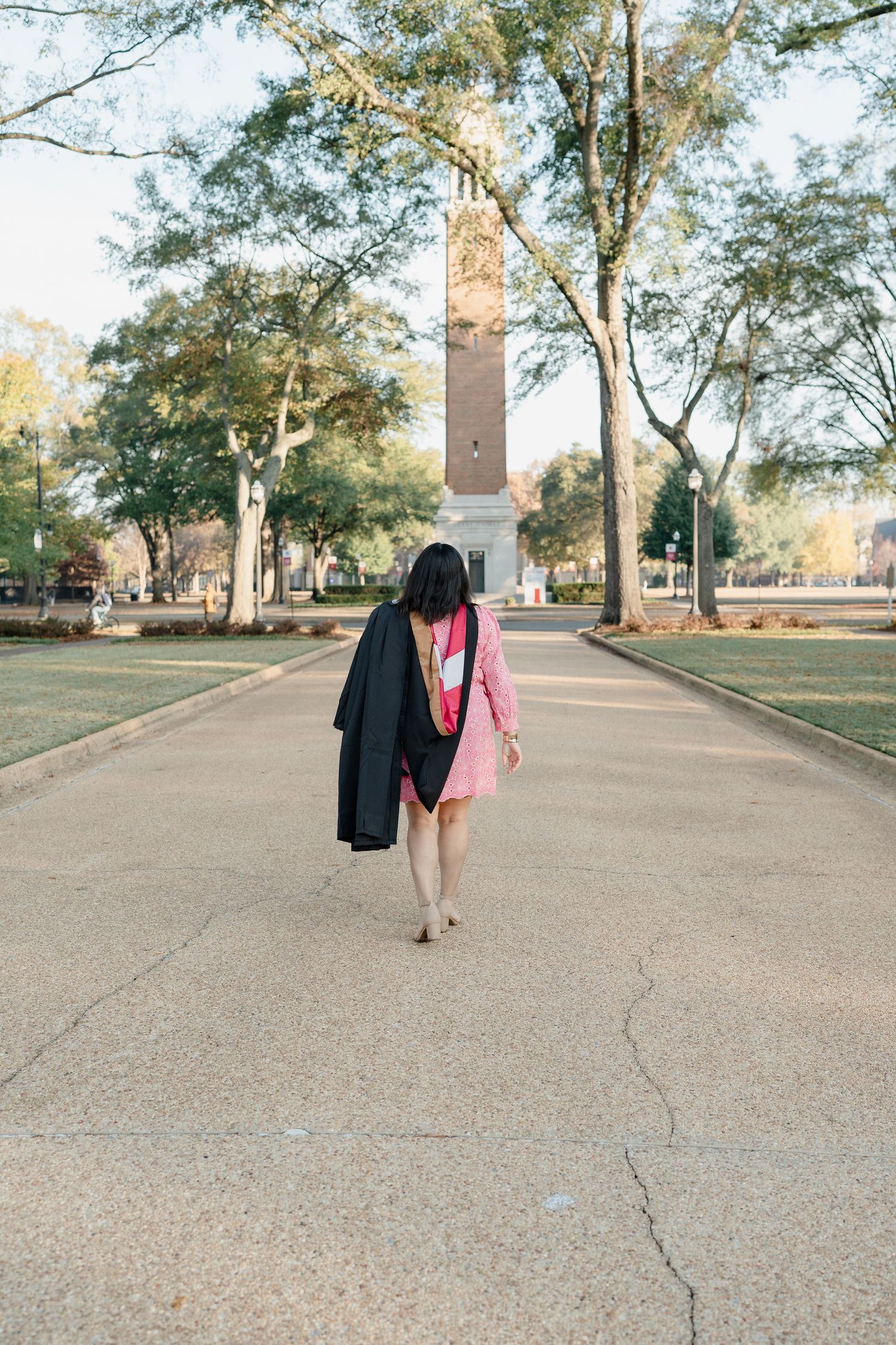 Graduate walking away from camera on campus path with graduation gown and monument in background.