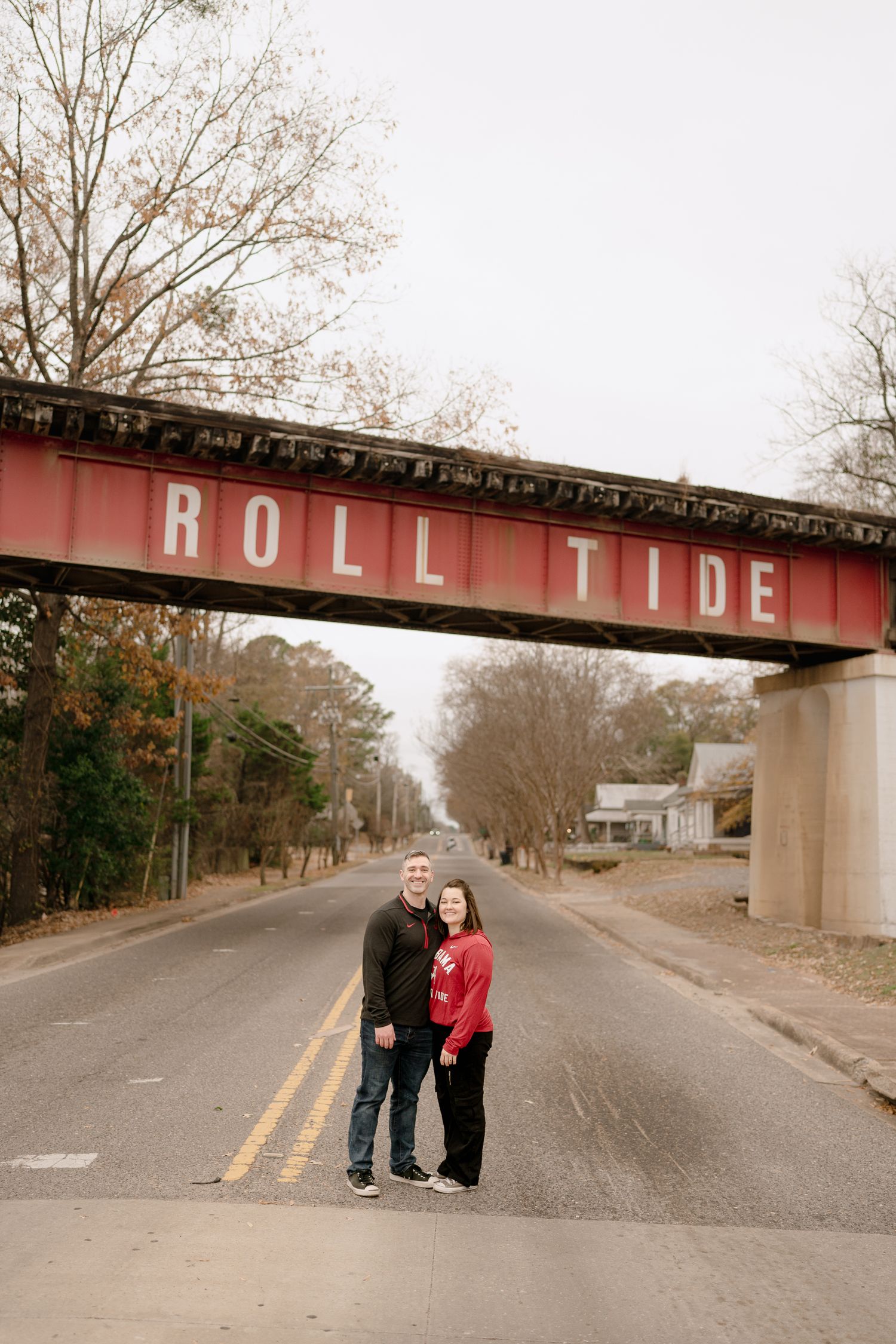 A couple stands beneath a red 'Roll Tide' bridge on an empty street in Alabama.