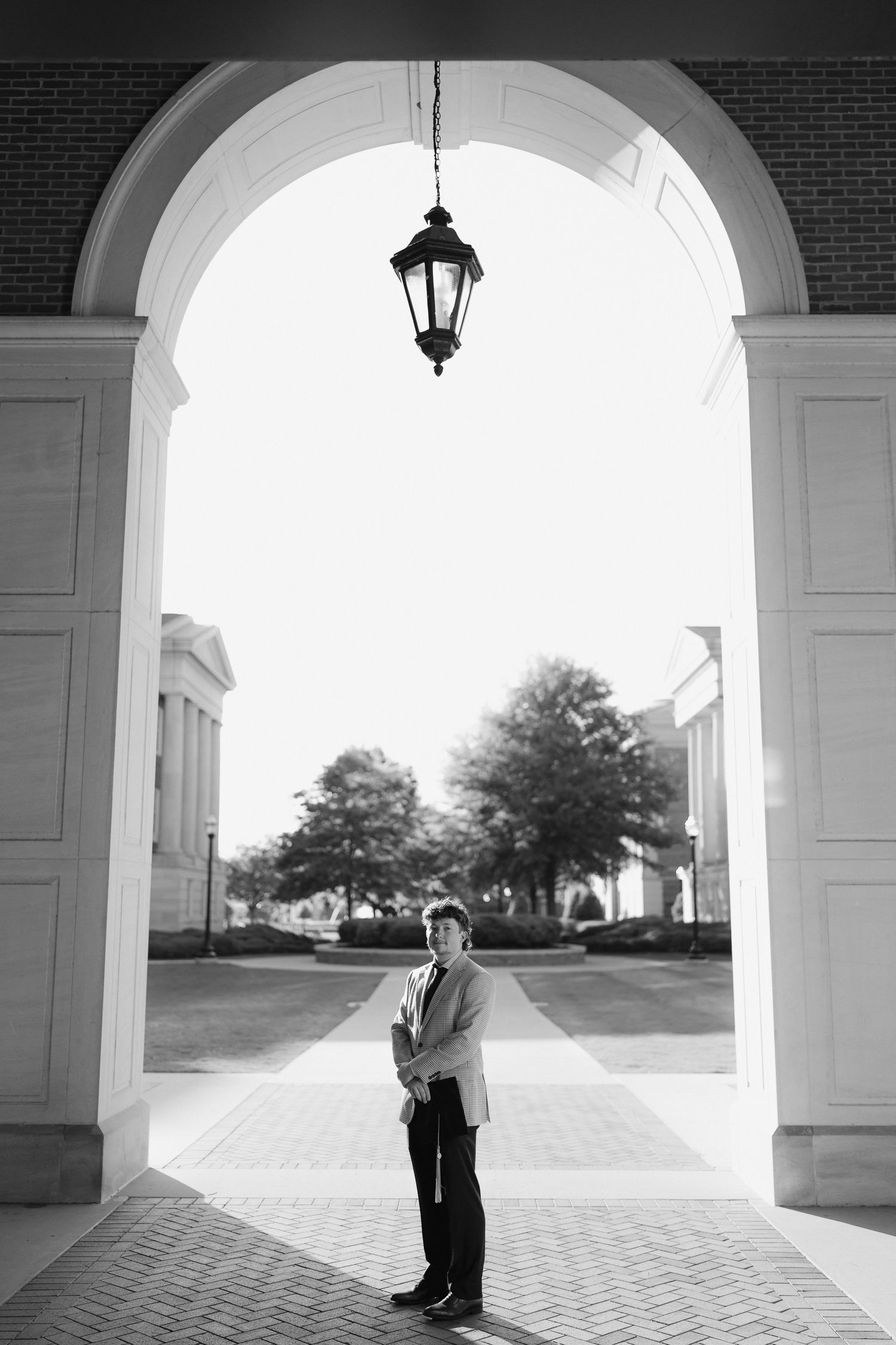 Black and white portrait of student standing under grand arched entrance with hanging lantern on campus.
