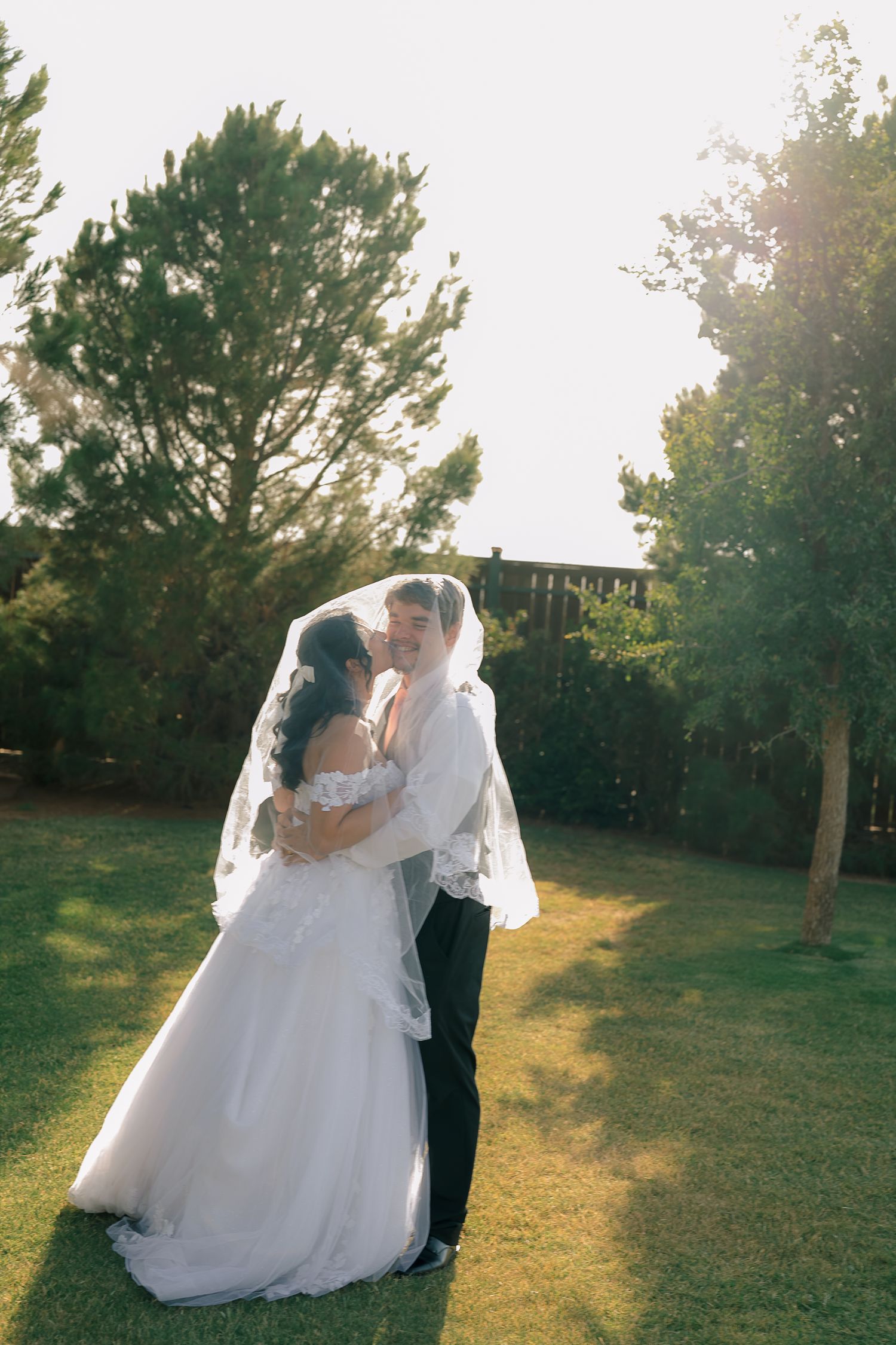 Bride and groom sharing a romantic kiss outdoors under a flowing veil, surrounded by lush green trees in golden sunlight.