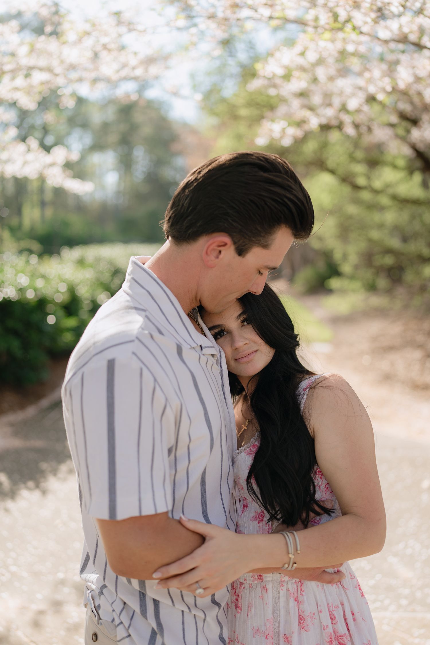 Couple embraces tenderly under flowering cherry blossom trees, woman resting head on mans chest in spring garden.
