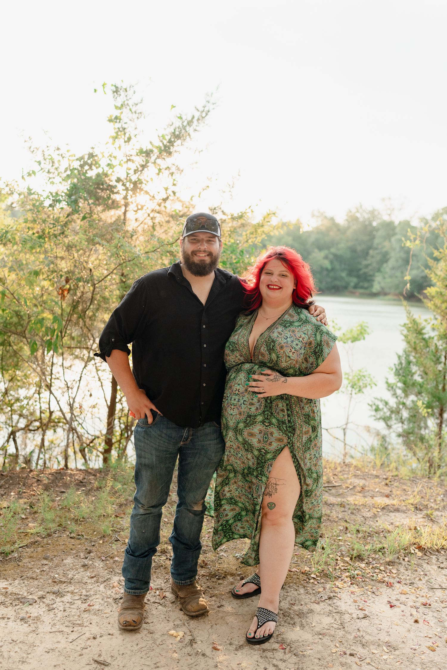 A couple poses playfully near a lake shore with trees and natural scenery in the background.