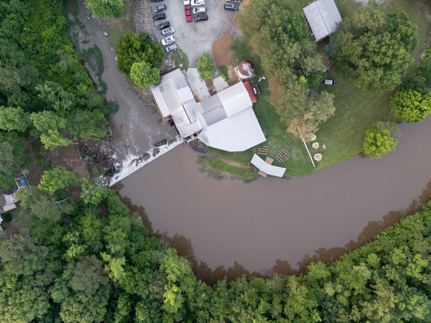 Aerial drone view of a riverside venue surrounded by lush green trees and a brown muddy river.