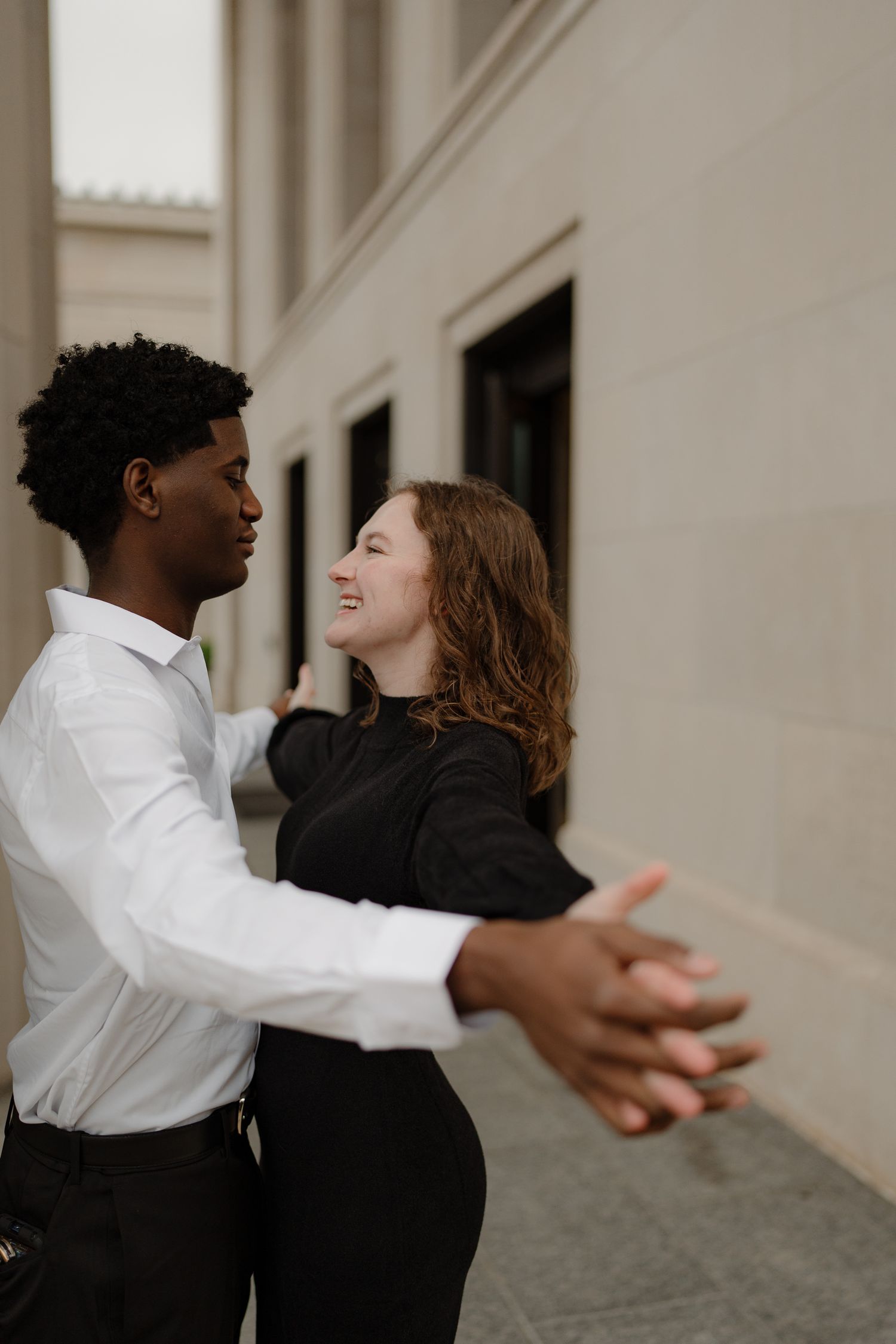 Smiling interracial couple dancing together outdoors, woman in black top, man in white dress shirt holding hands.