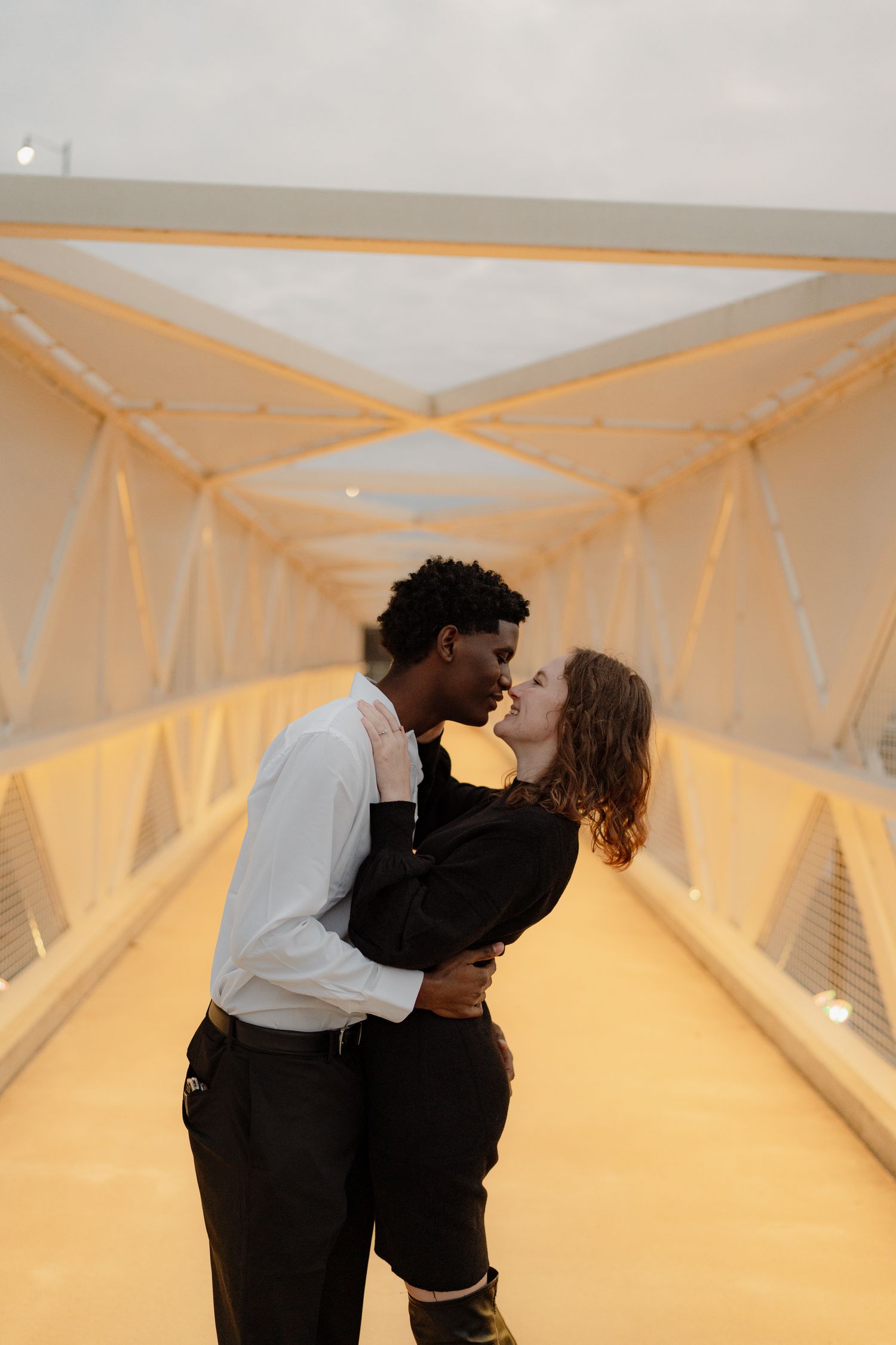 An interracial couple shares a tender almost-kiss on a warmly lit enclosed bridge walkway.
