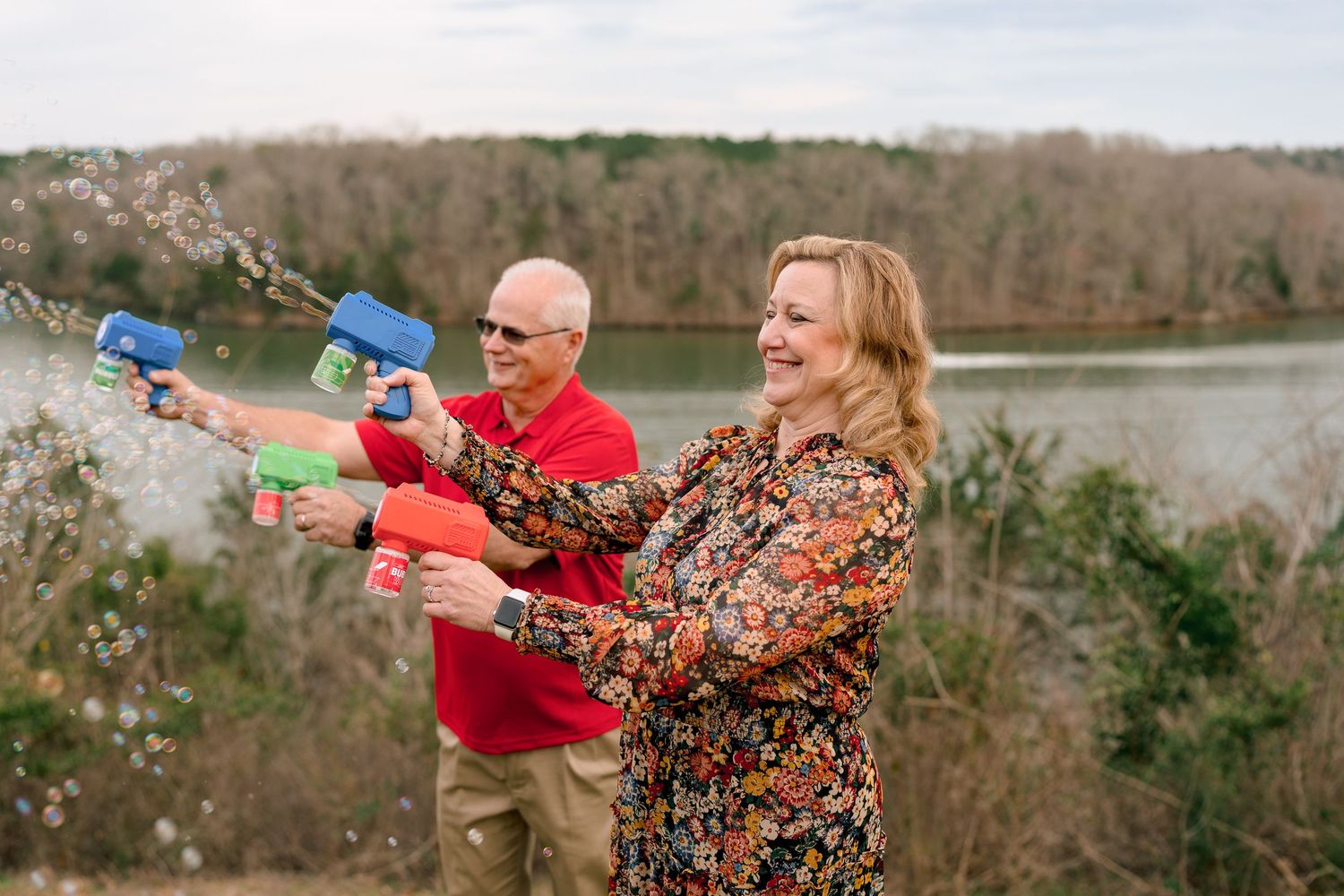 Playful couple shooting water guns together near a lake, laughing and having fun outdoors.