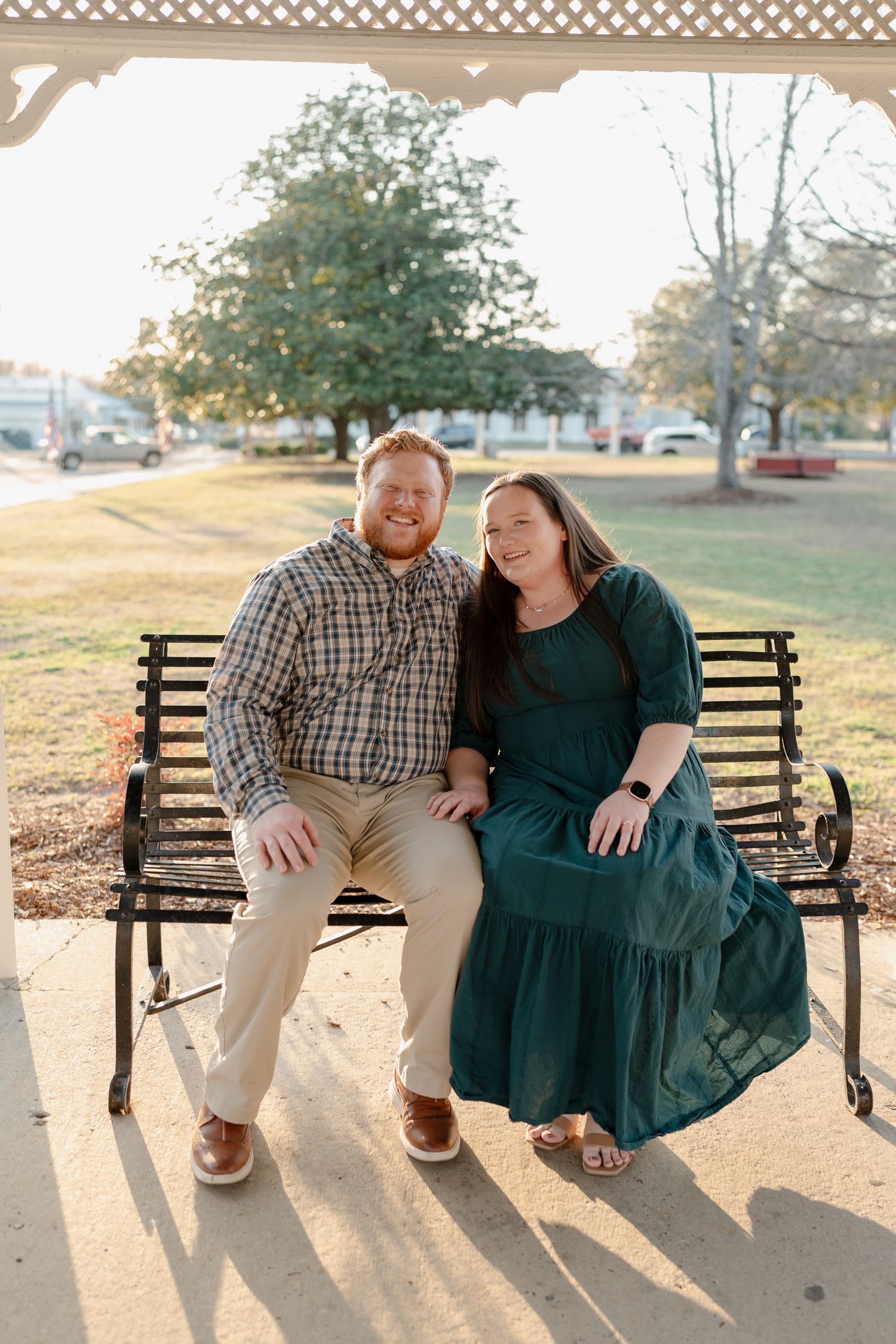 Happy couple sitting on a black iron bench outdoors, man in plaid shirt and woman in emerald green maxi dress, smiling.