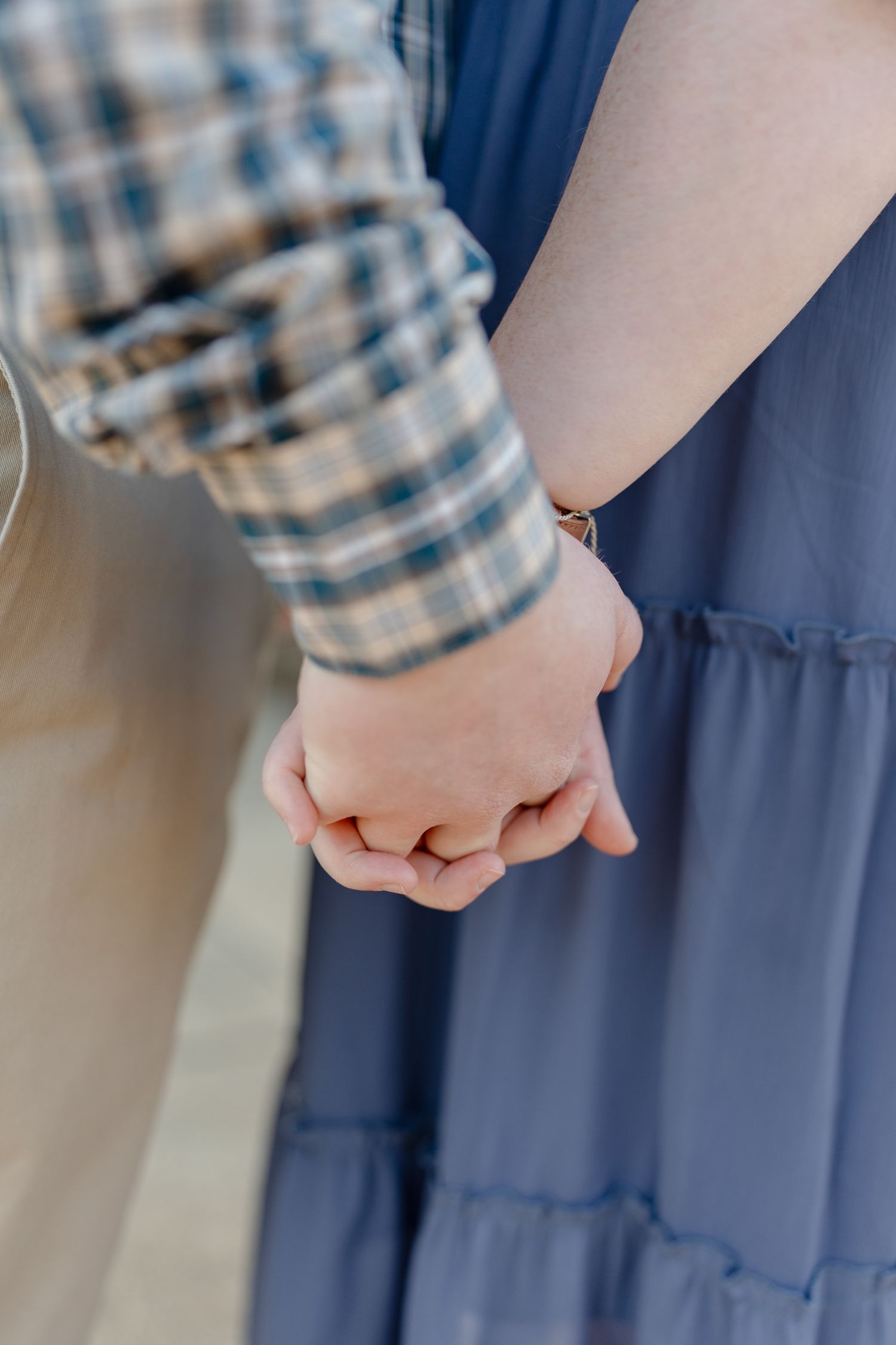 Close-up of a couple holding hands, woman in blue tiered dress with bracelet, man in plaid shirt.