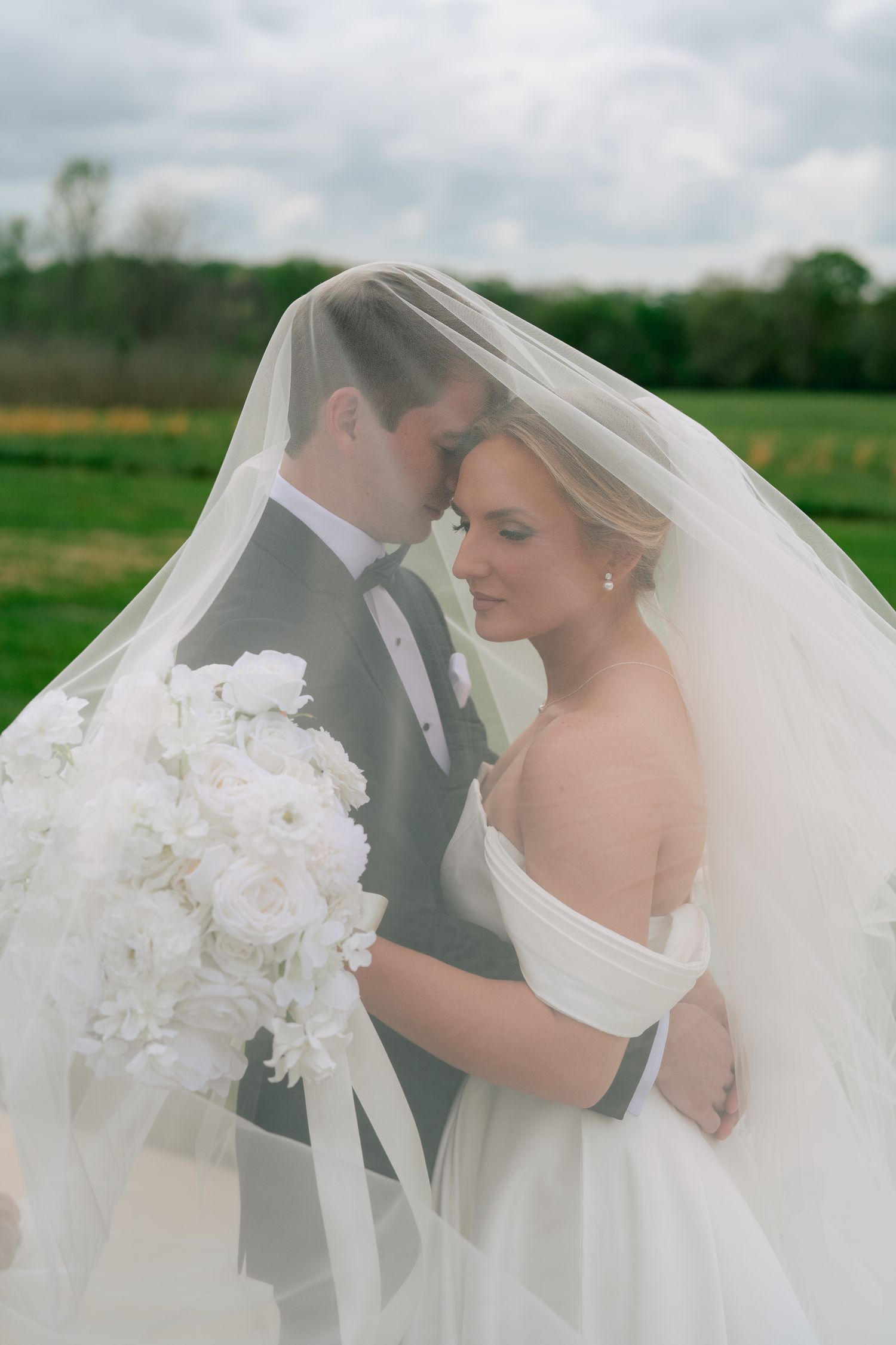 Bride and groom share an intimate moment under a flowing white veil, with a lush green field backdrop on their wedding day.