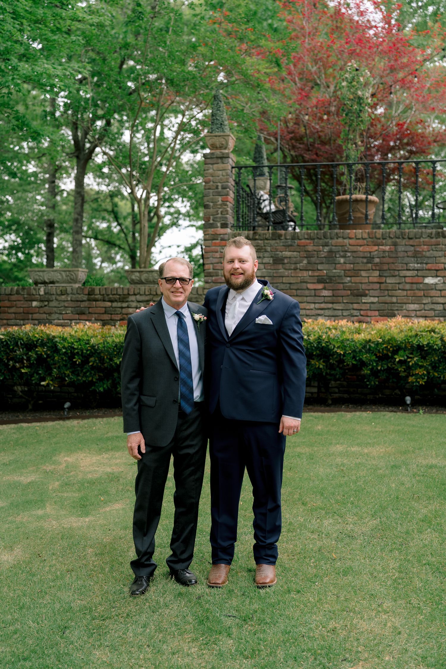 Two men in suits pose together outdoors at a wedding venue with brick wall and lush greenery.